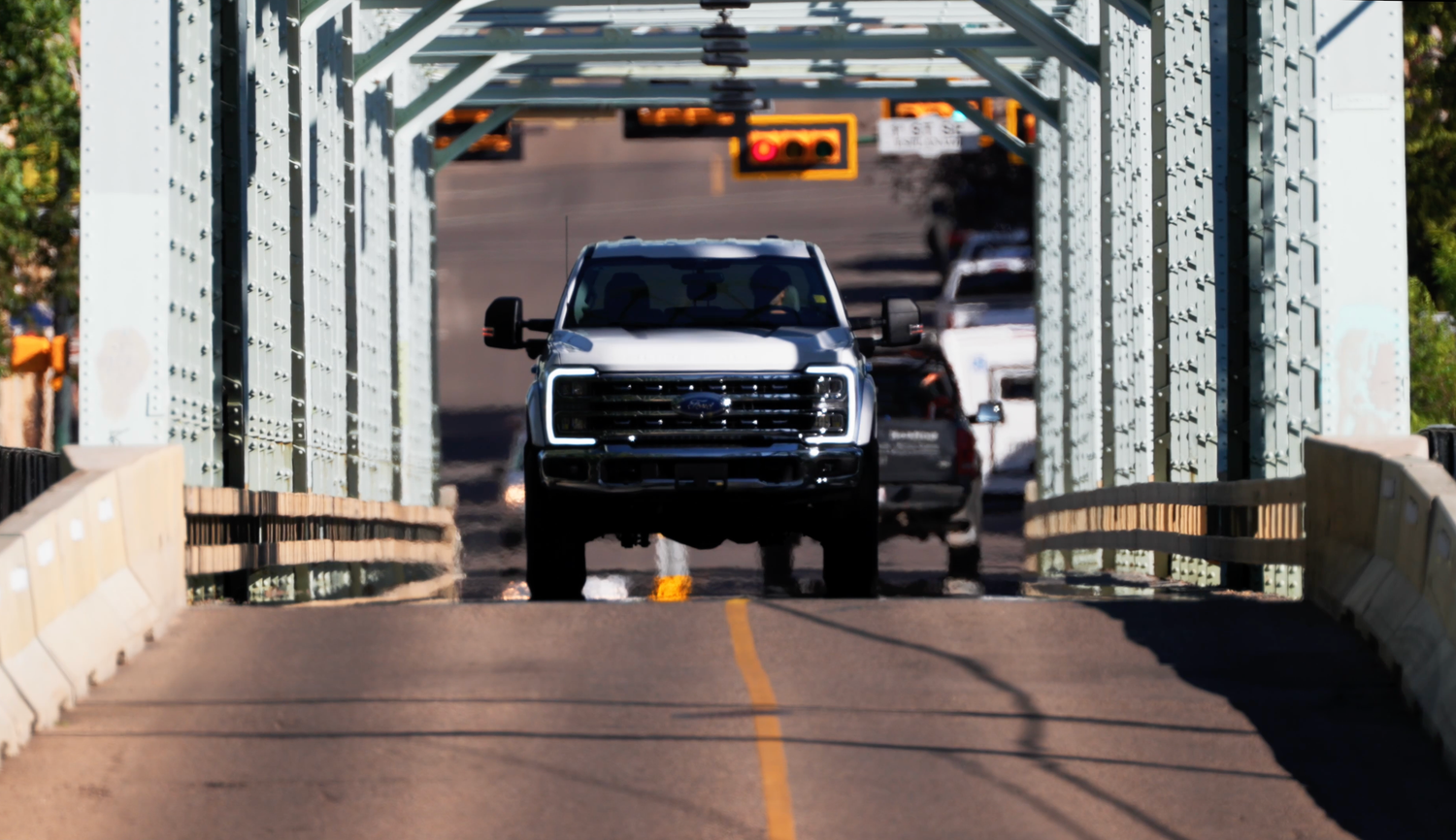 White pickup truck driving over a steel truss bridge with traffic visible in background on a sunny day.
