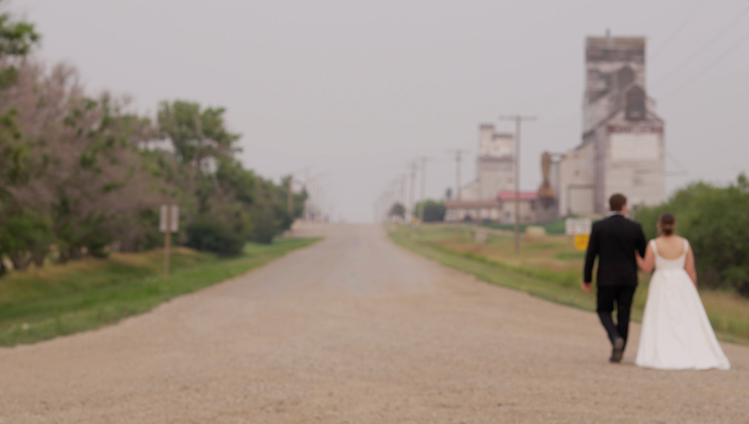 Bride and groom walking hand in hand down a rural road with grain elevators in the misty background of Fox Valley.