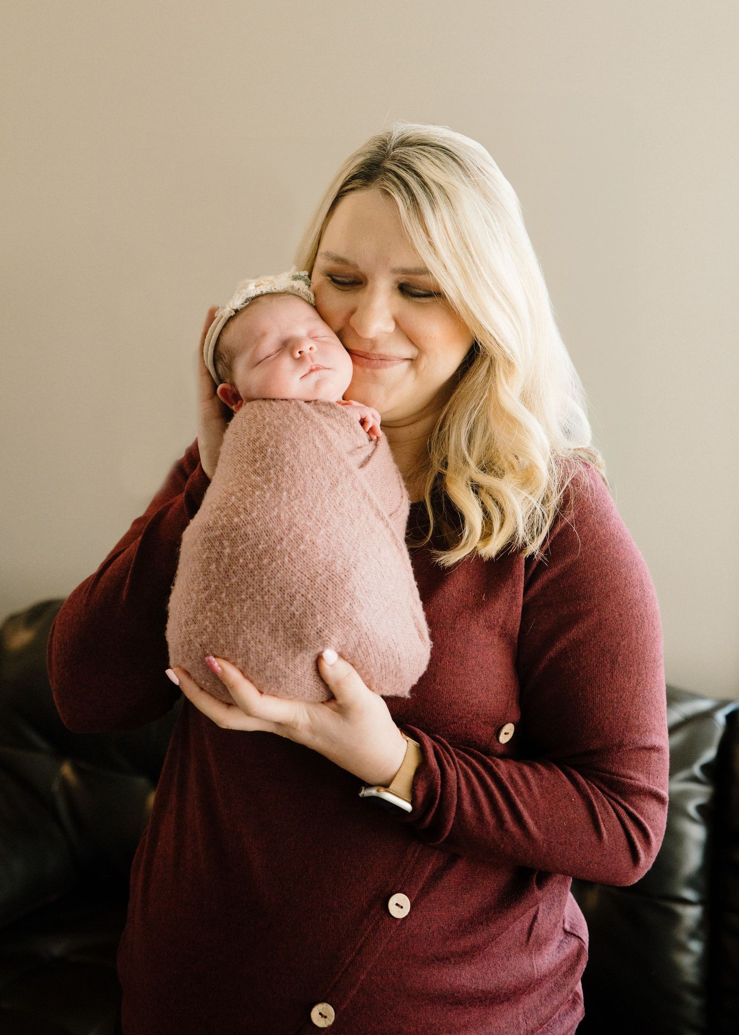Smiling blonde woman in burgundy dress holding sleeping newborn swaddled in pink blanket.
