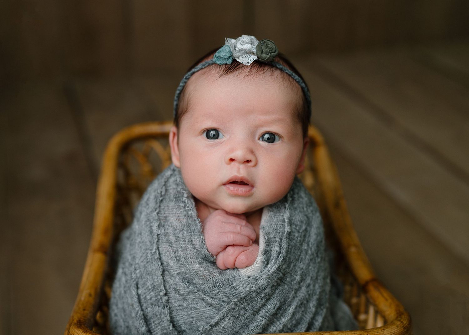 Alert newborn baby swaddled in gray wrap with floral headband sitting in wicker basket on wood floor.