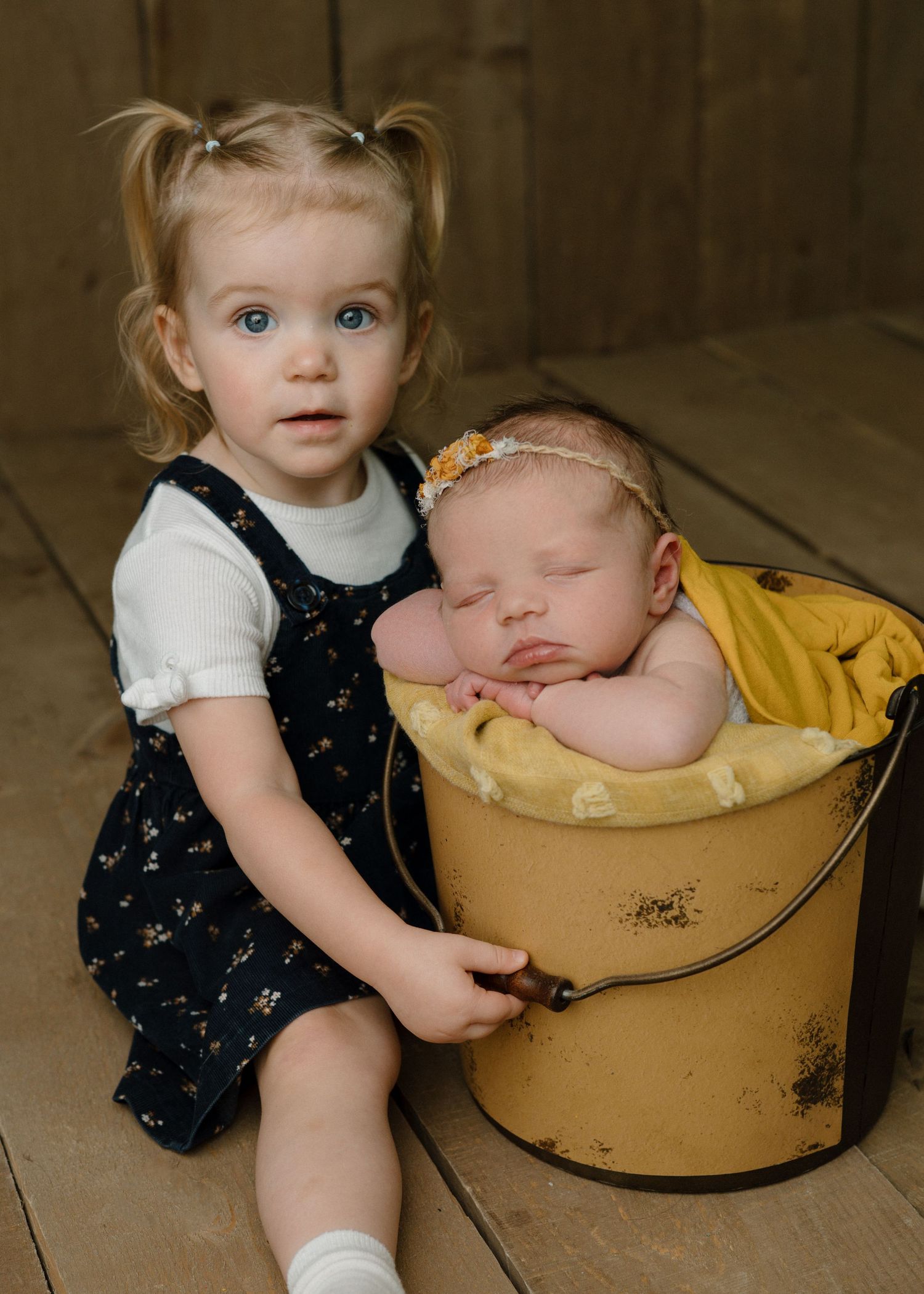Toddler girl with pigtails sitting beside a vintage yellow bucket holding a sleeping newborn baby wrapped in yellow.