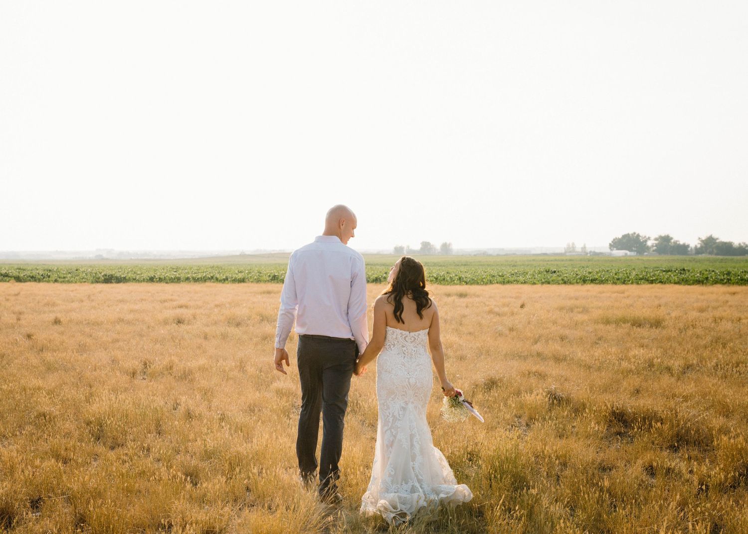 Bride and groom walk hand in hand through a golden wheat field during sunset wedding portraits.