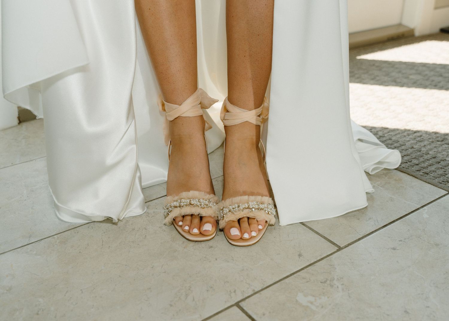 Close-up of a bride wearing elegant beige strappy heeled sandals with fringe detail, standing on stone tiles in a white gown.