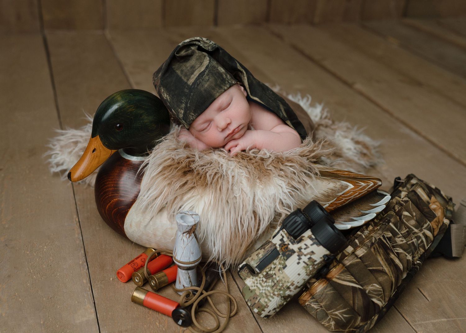 Newborn baby sleeping in camouflage hat surrounded by duck hunting props including decoy and shotgun shells.