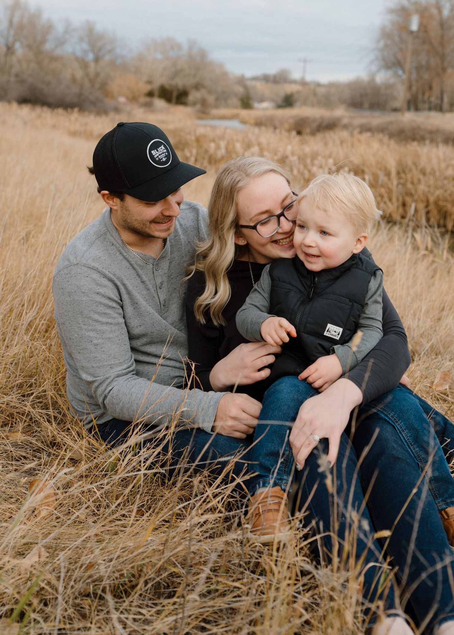 Young family of three sitting together in a golden dry grass field during autumn season.