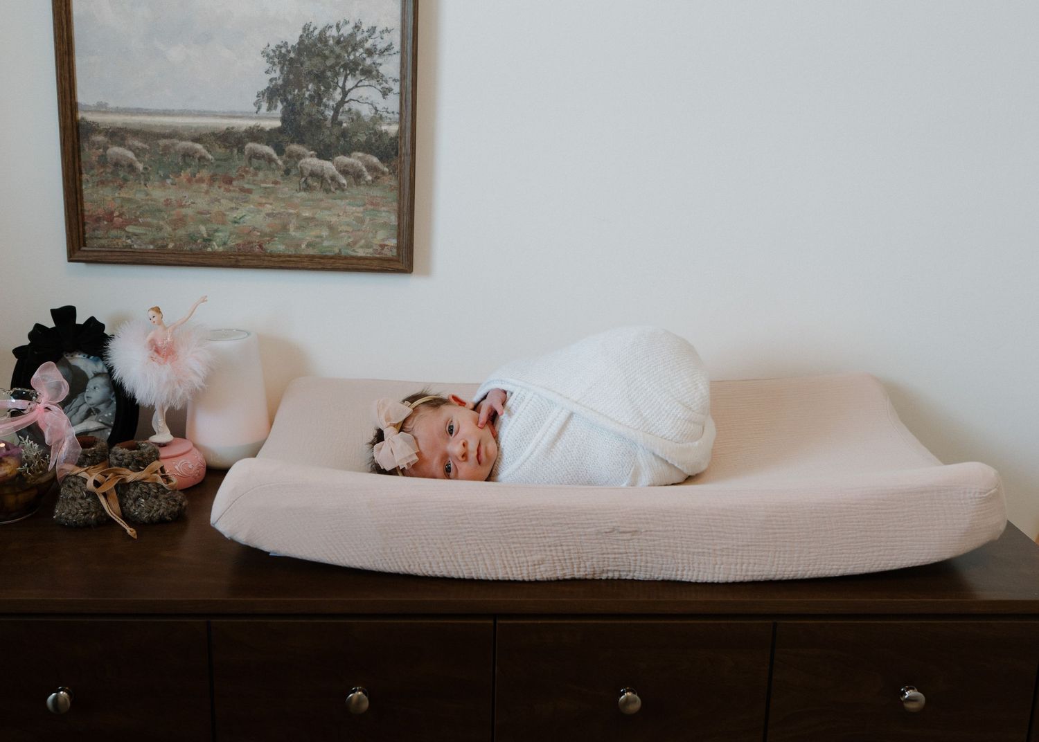 Swaddled newborn sleeping peacefully on changing pad beside vintage sheep painting.