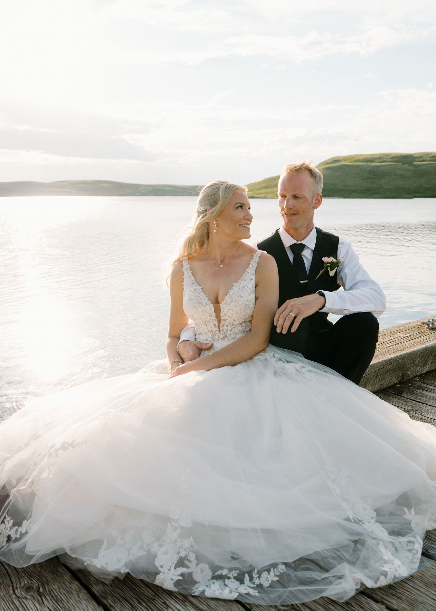 Bride and groom sitting on a dock by a serene lake, bride in a lace ballgown, groom in a black vest.