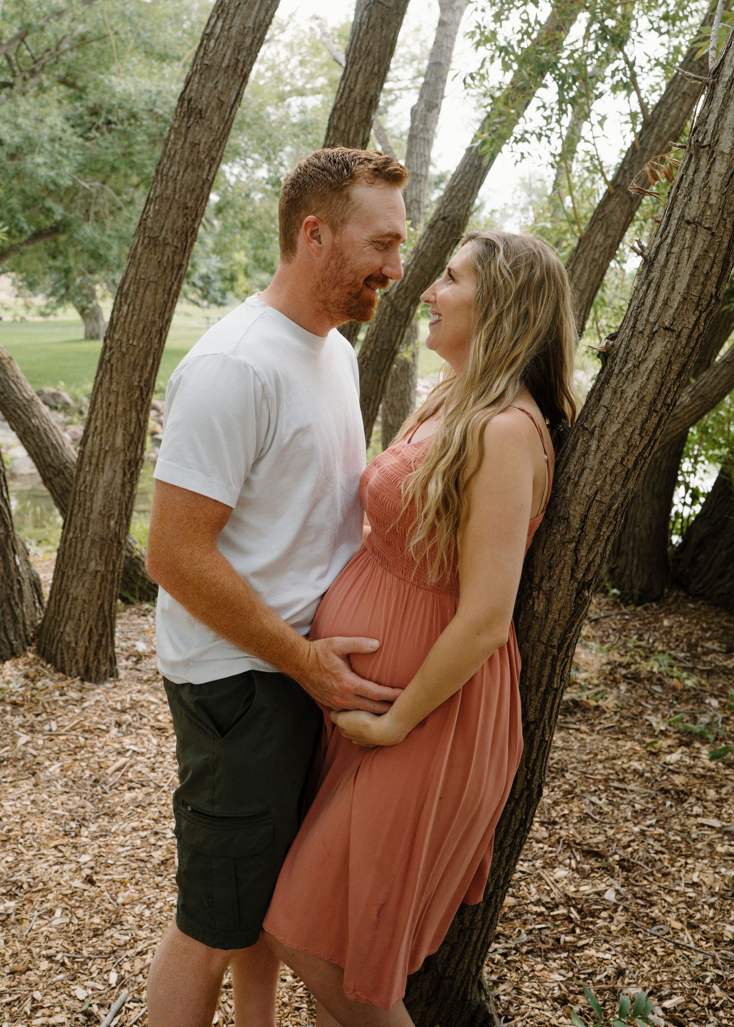Couple embracing near trees, pregnant woman in coral dress smiling at her partner.