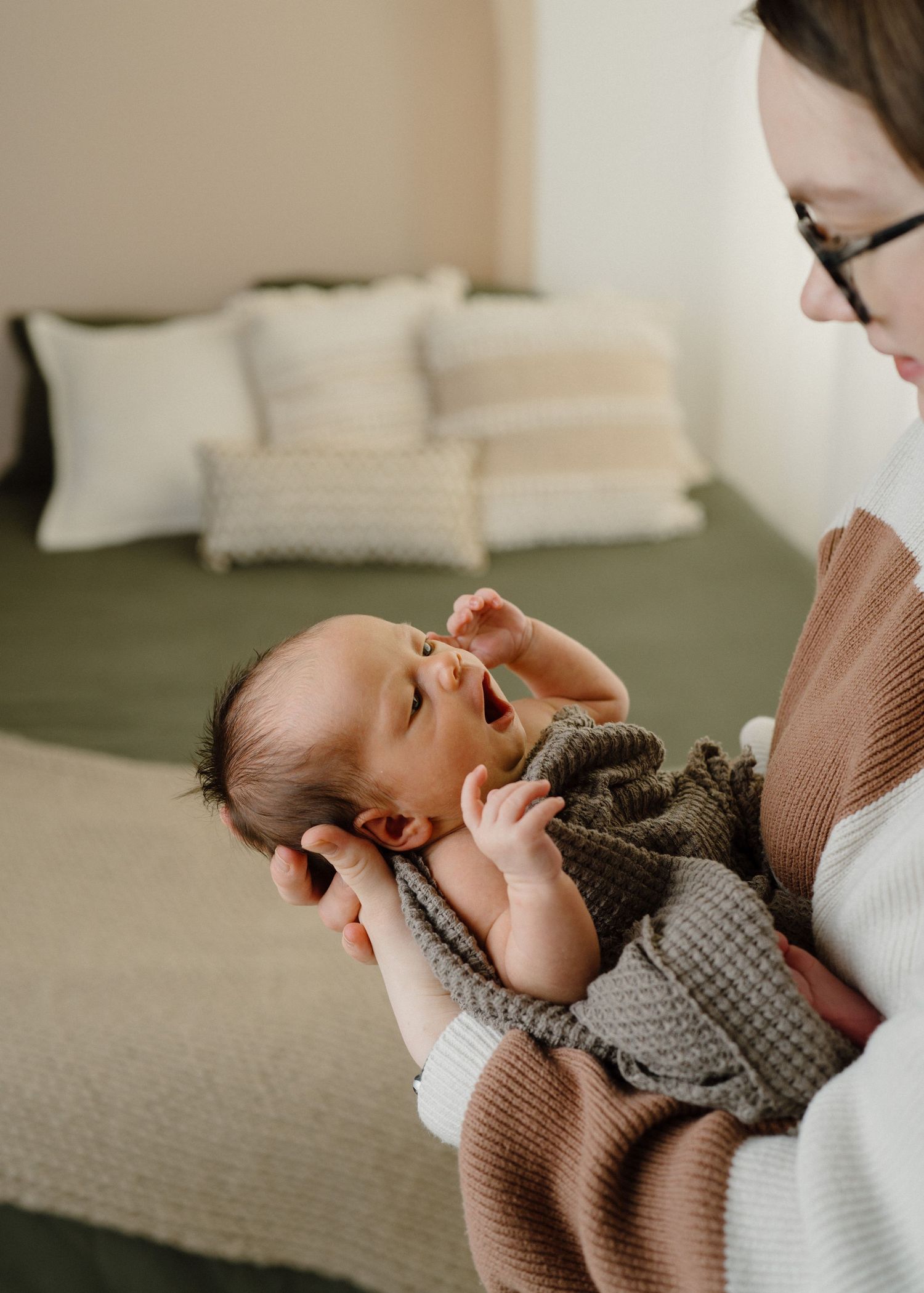 Mother in brown sweater holding alert newborn baby wrapped in olive knit blanket indoors.