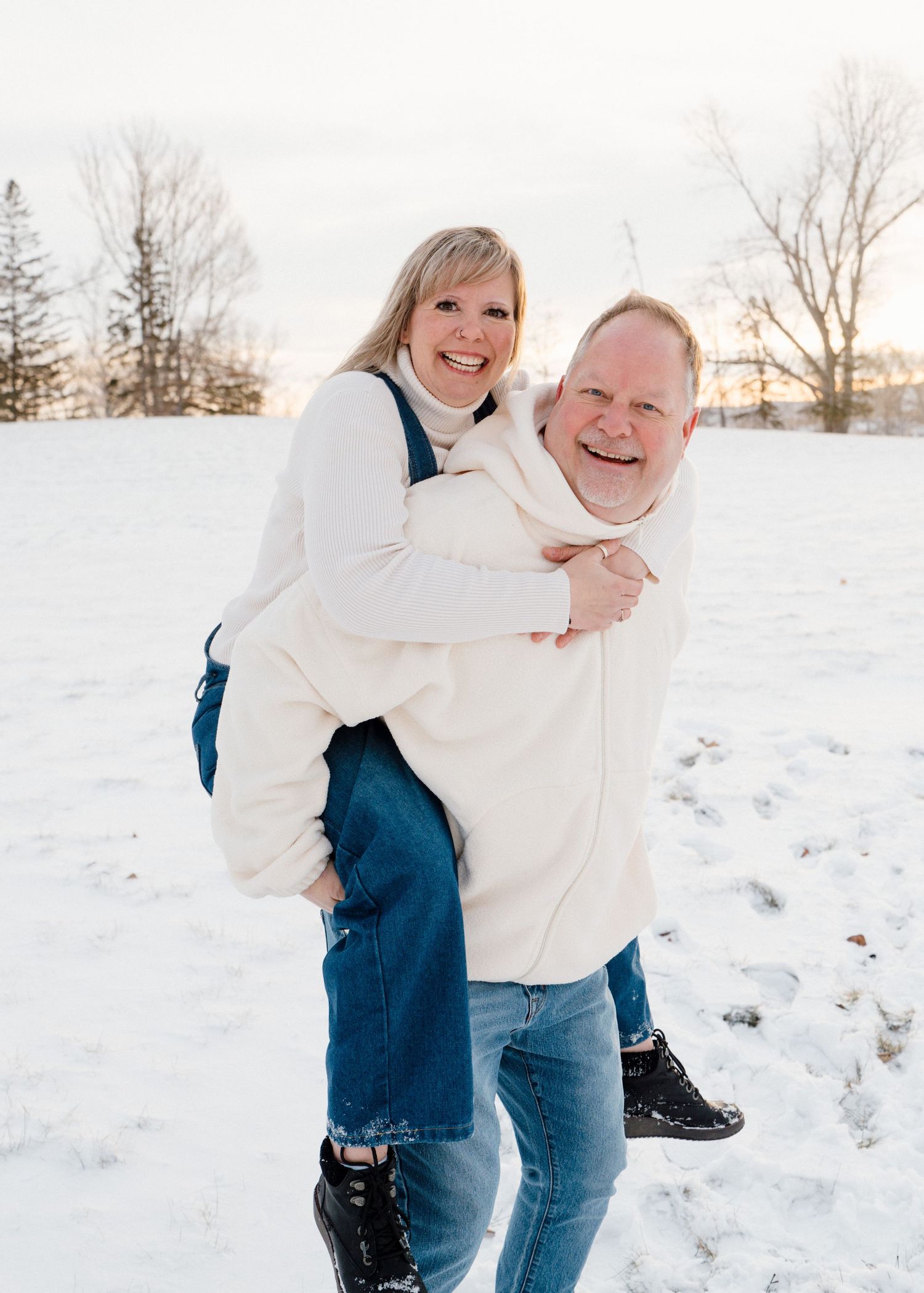 Happy couple laughing as man gives woman a piggyback ride in snowy winter setting.