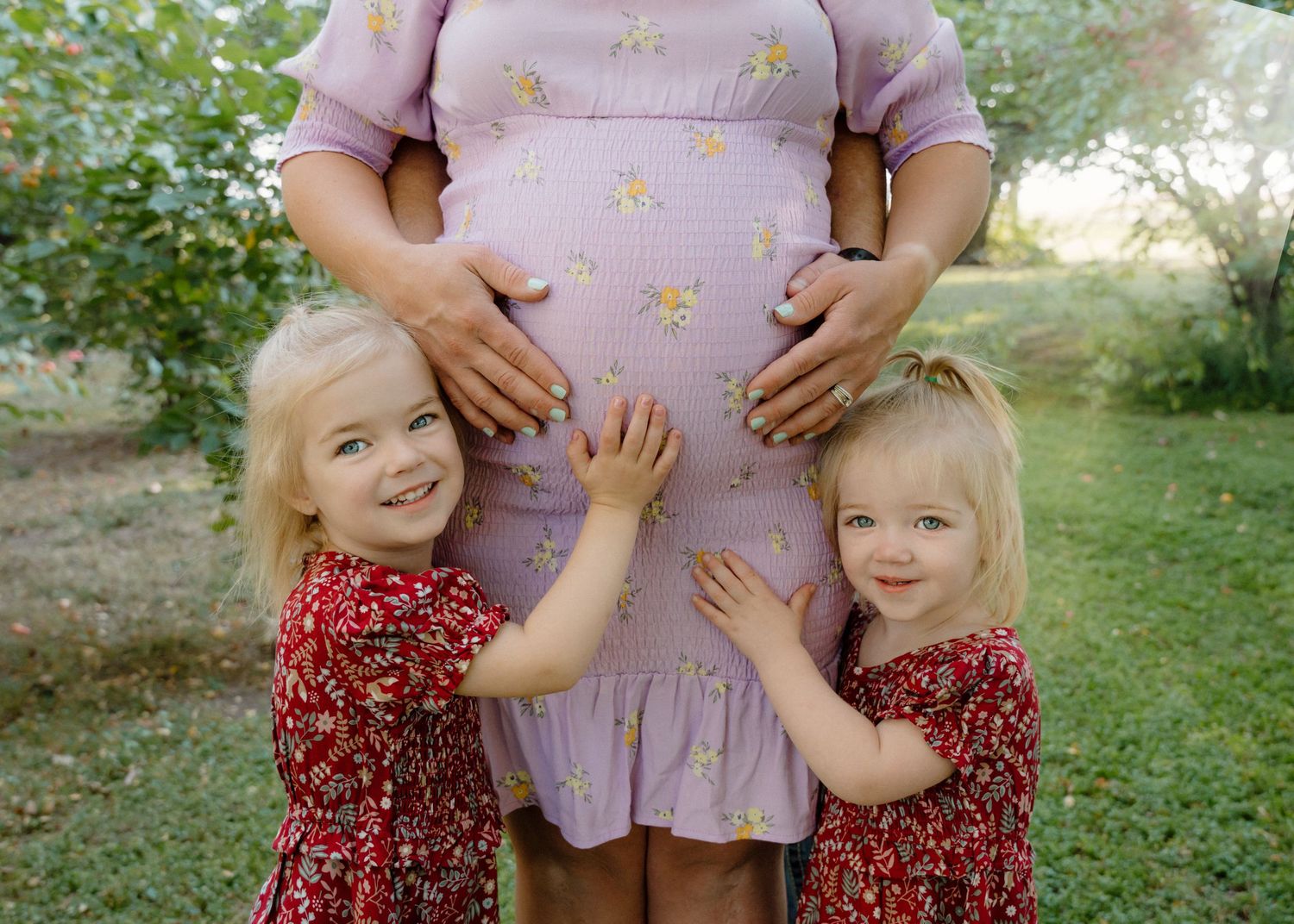 Two young sisters in red floral dresses touching pregnant mothers belly in garden setting.