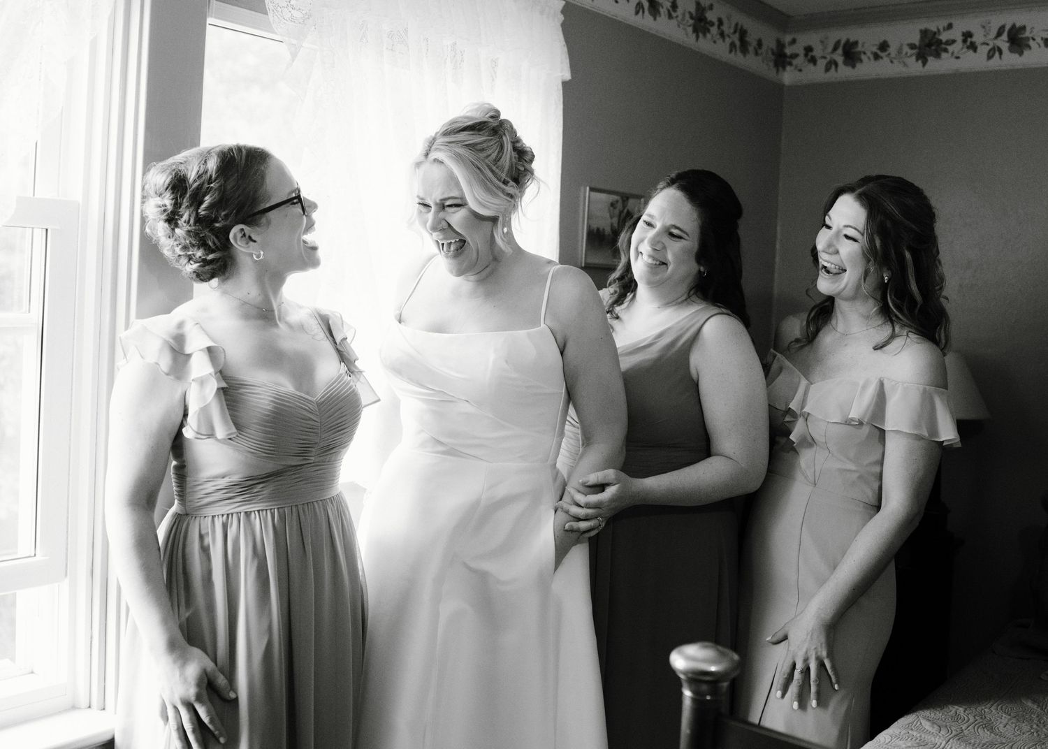 Black and white photo of a bride laughing with three bridesmaids while getting ready indoors.