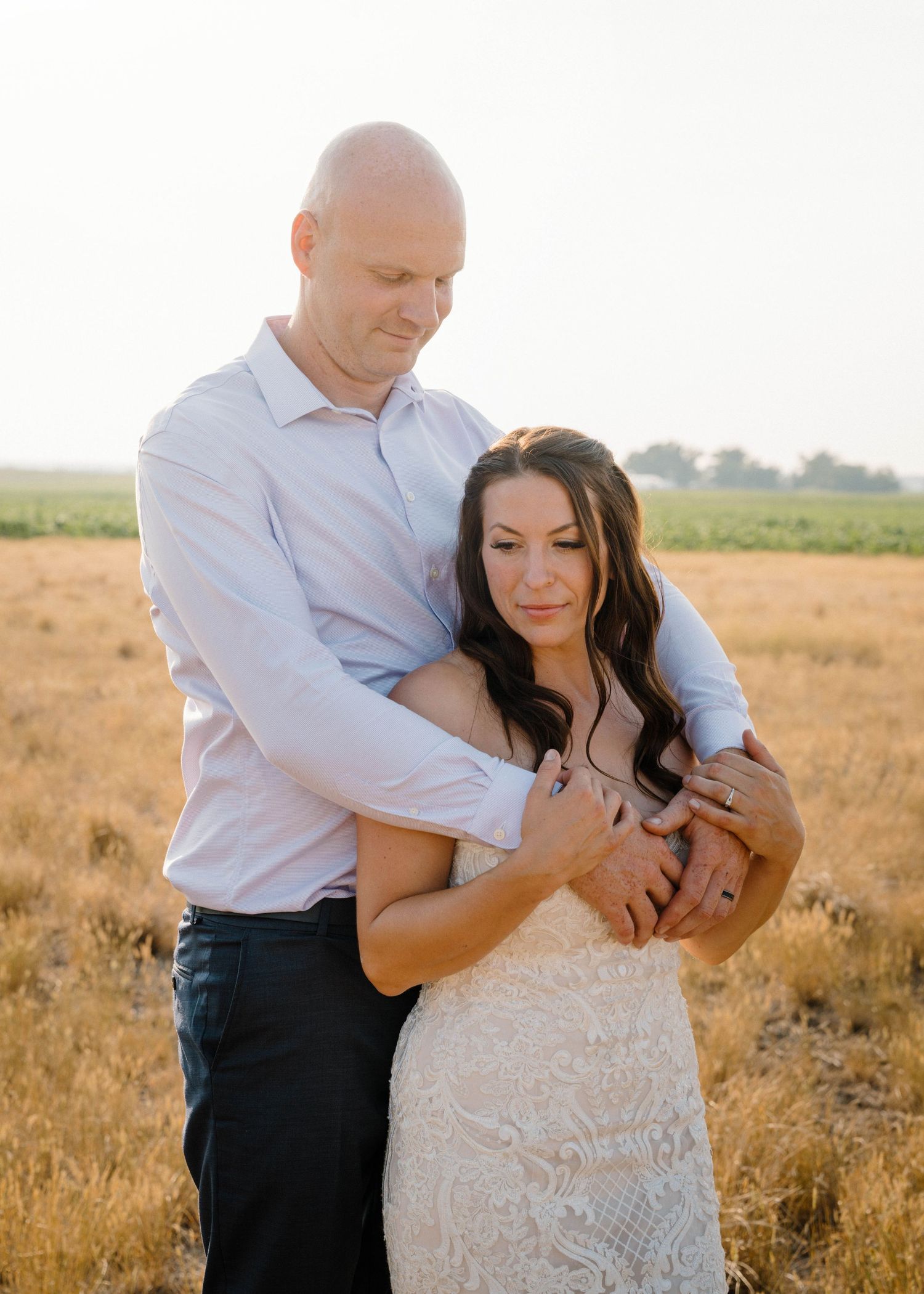 Groom embraces bride from behind in a warm sunlit field during romantic outdoor wedding portraits.