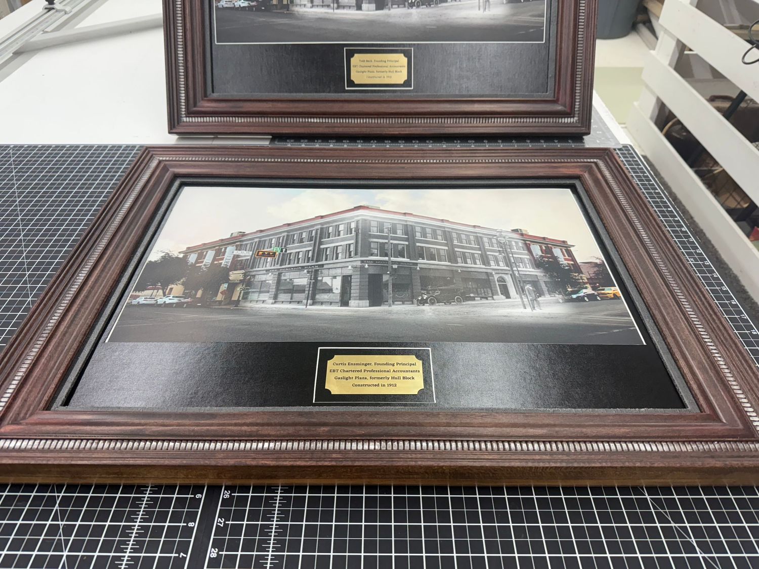 Two ornate wood framed vintage building photographs with gold engraved plaques on workbench.