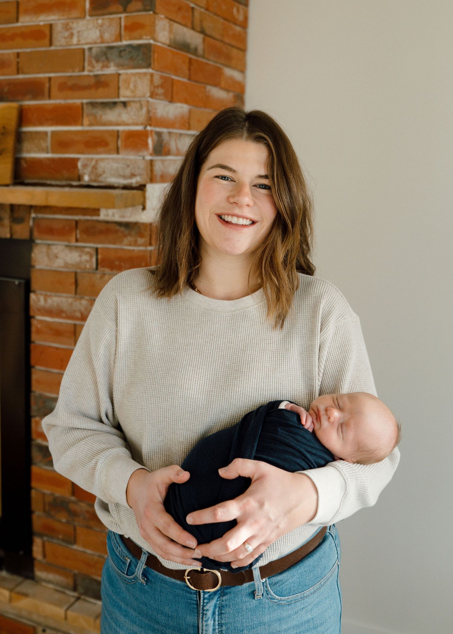 Smiling brunette woman holding swaddled newborn in navy wrap standing by brick fireplace.