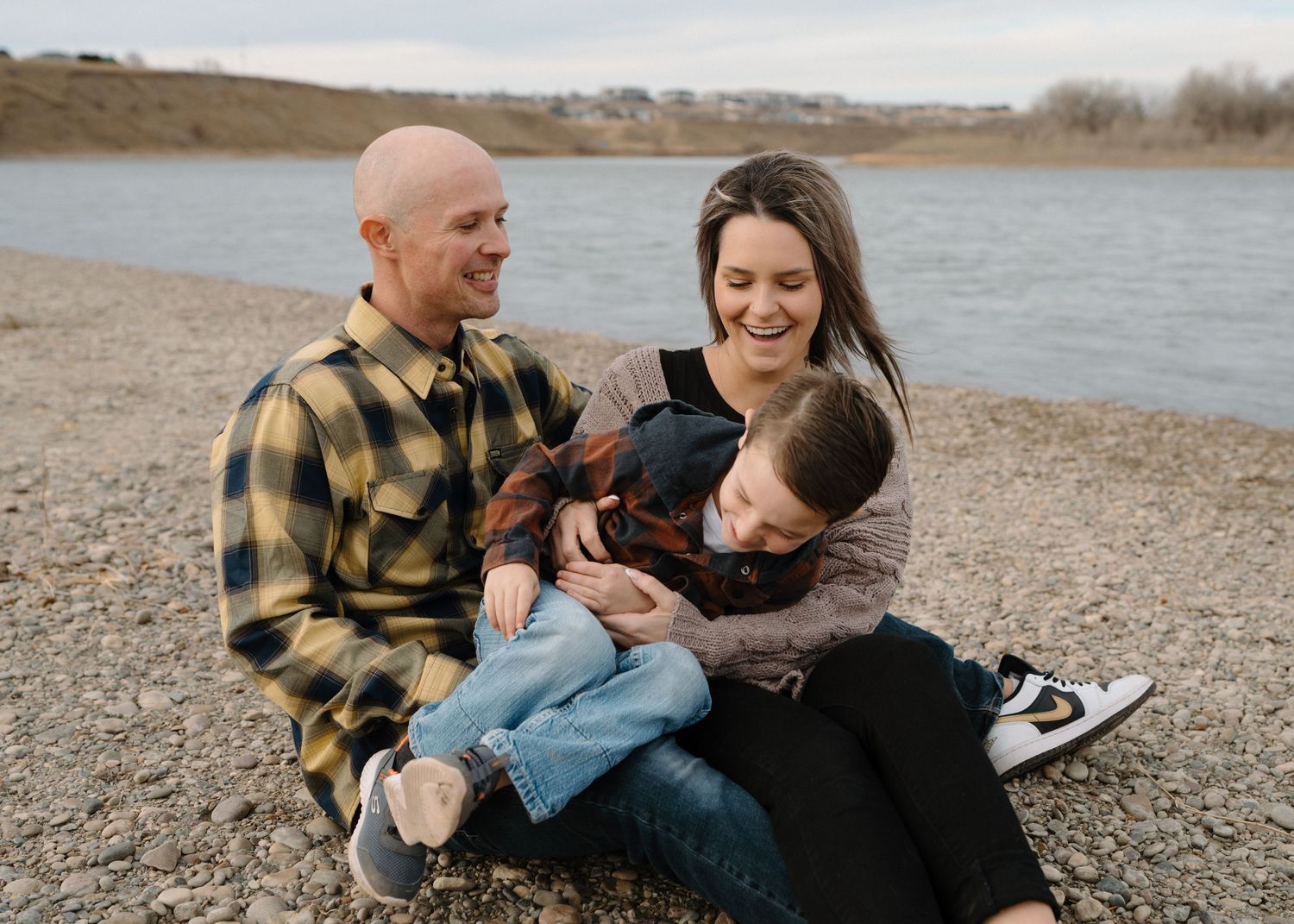 Joyful family sitting by a riverside, parents laughing with young child on mom's lap.