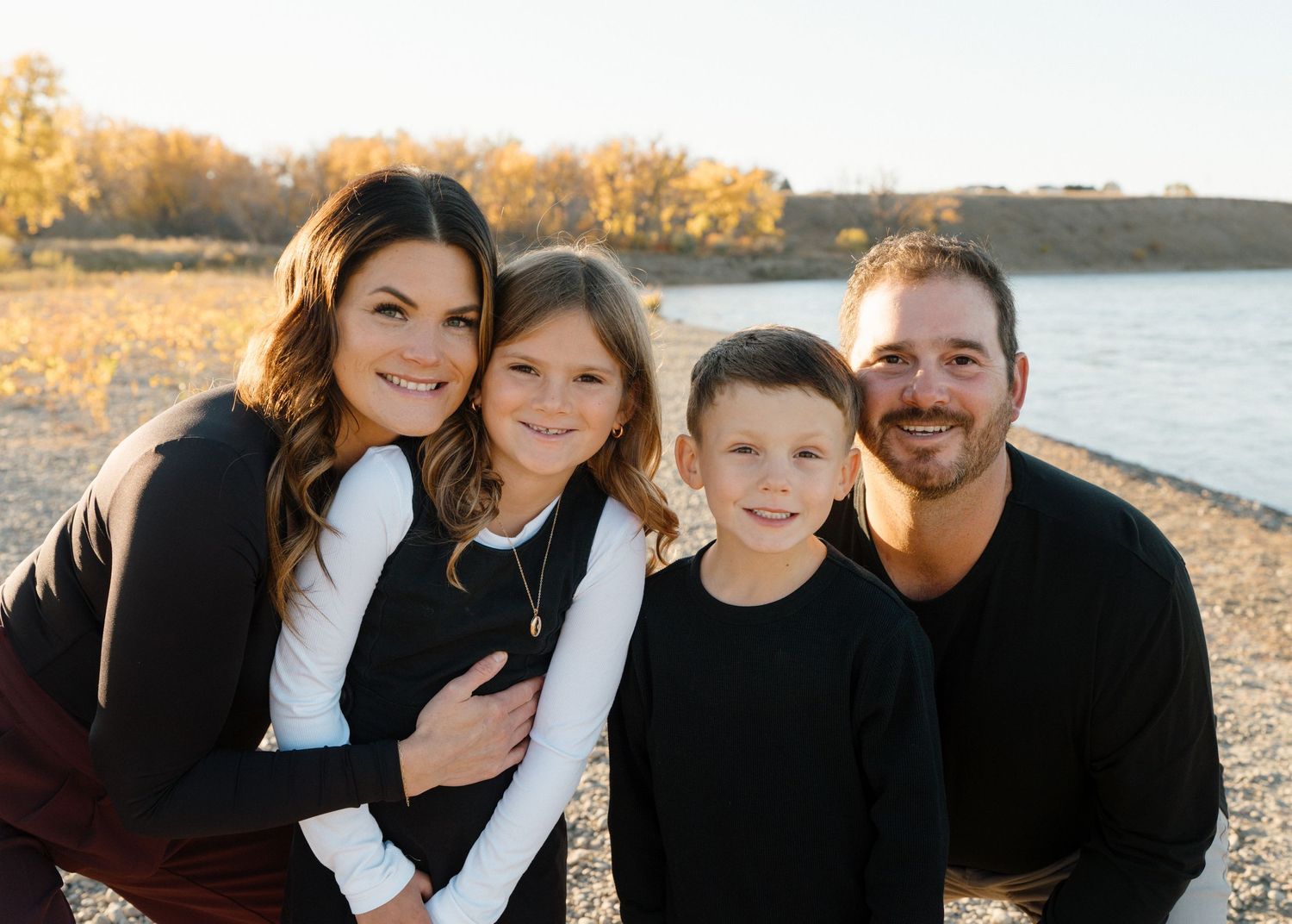 Smiling family of four enjoys a sunny autumn day together near a river with colorful fall foliage.