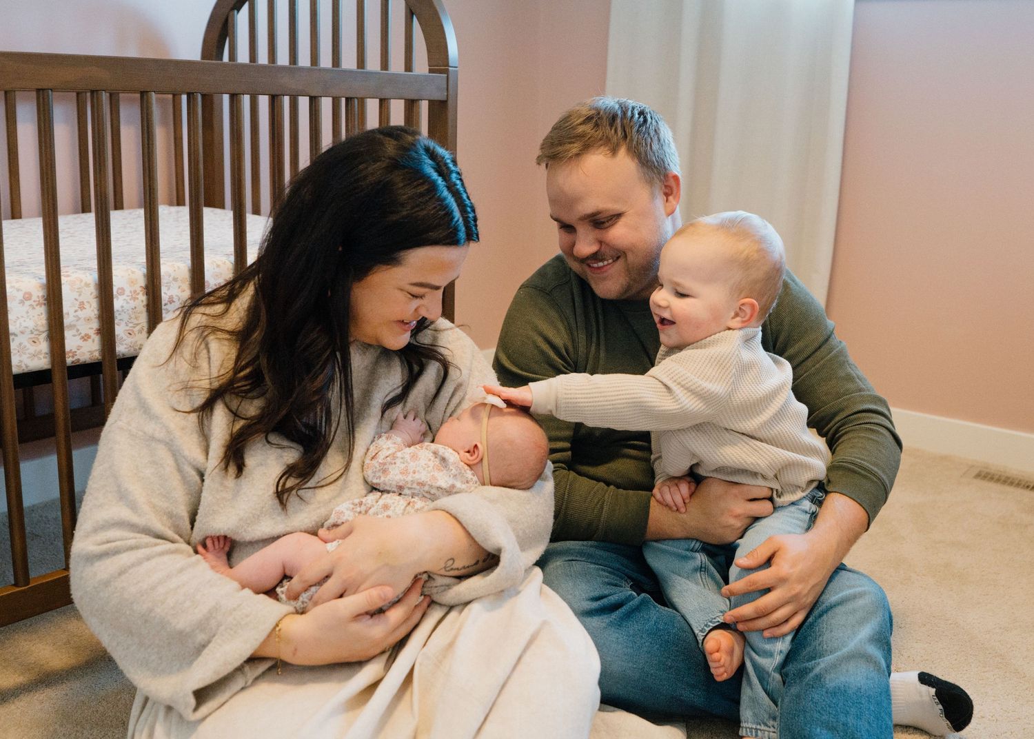 Happy family of four with mother holding newborn baby while father holds smiling older baby in a nursery.