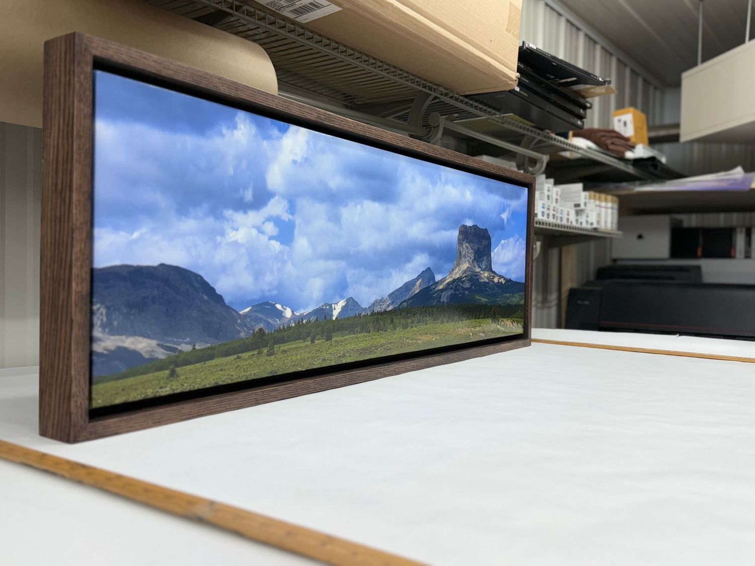 Panoramic landscape photo in dark wood frame showing mountains and green meadow under blue sky.