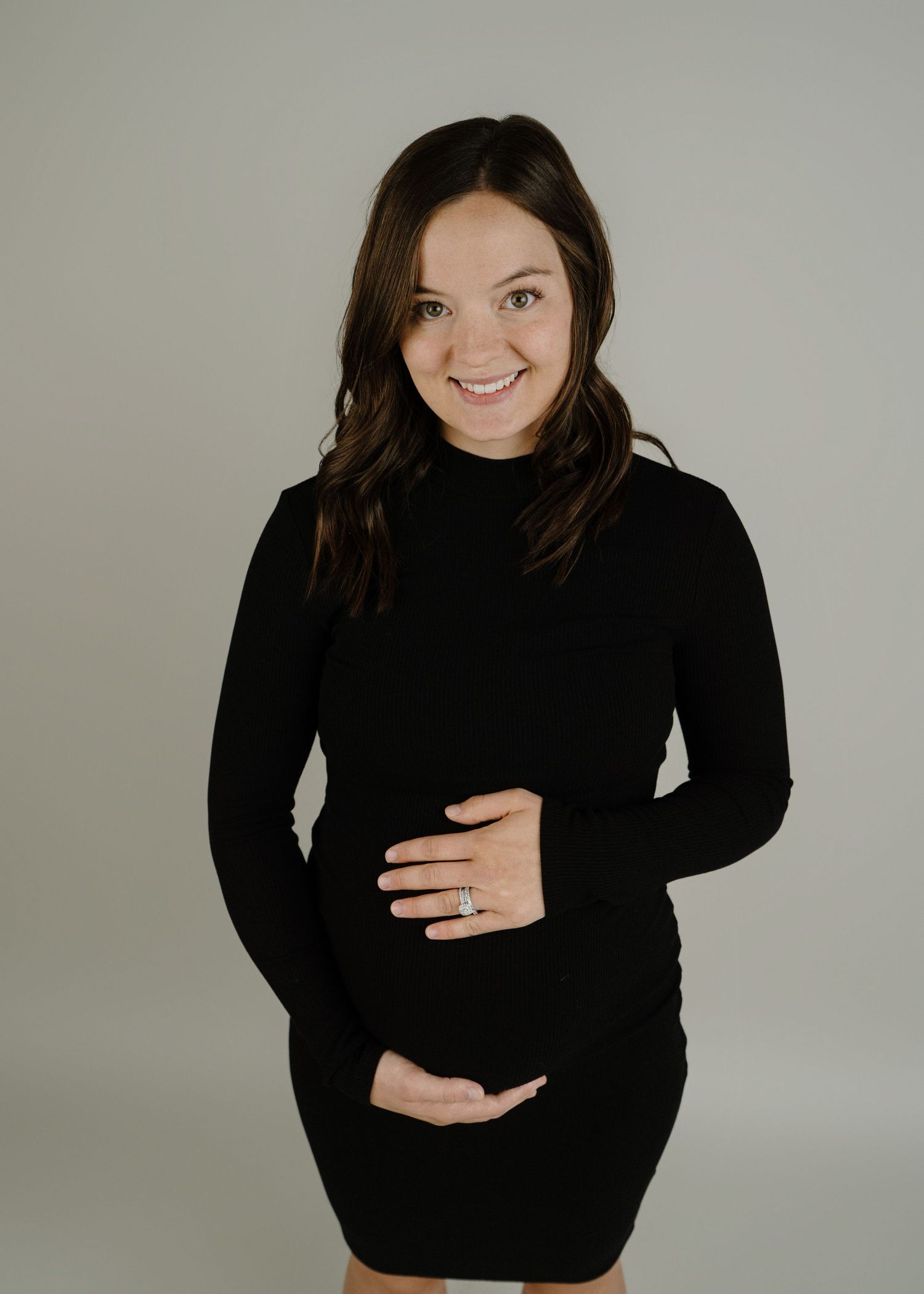 Smiling pregnant woman in a black turtleneck dress cradles her baby bump in a studio portrait.