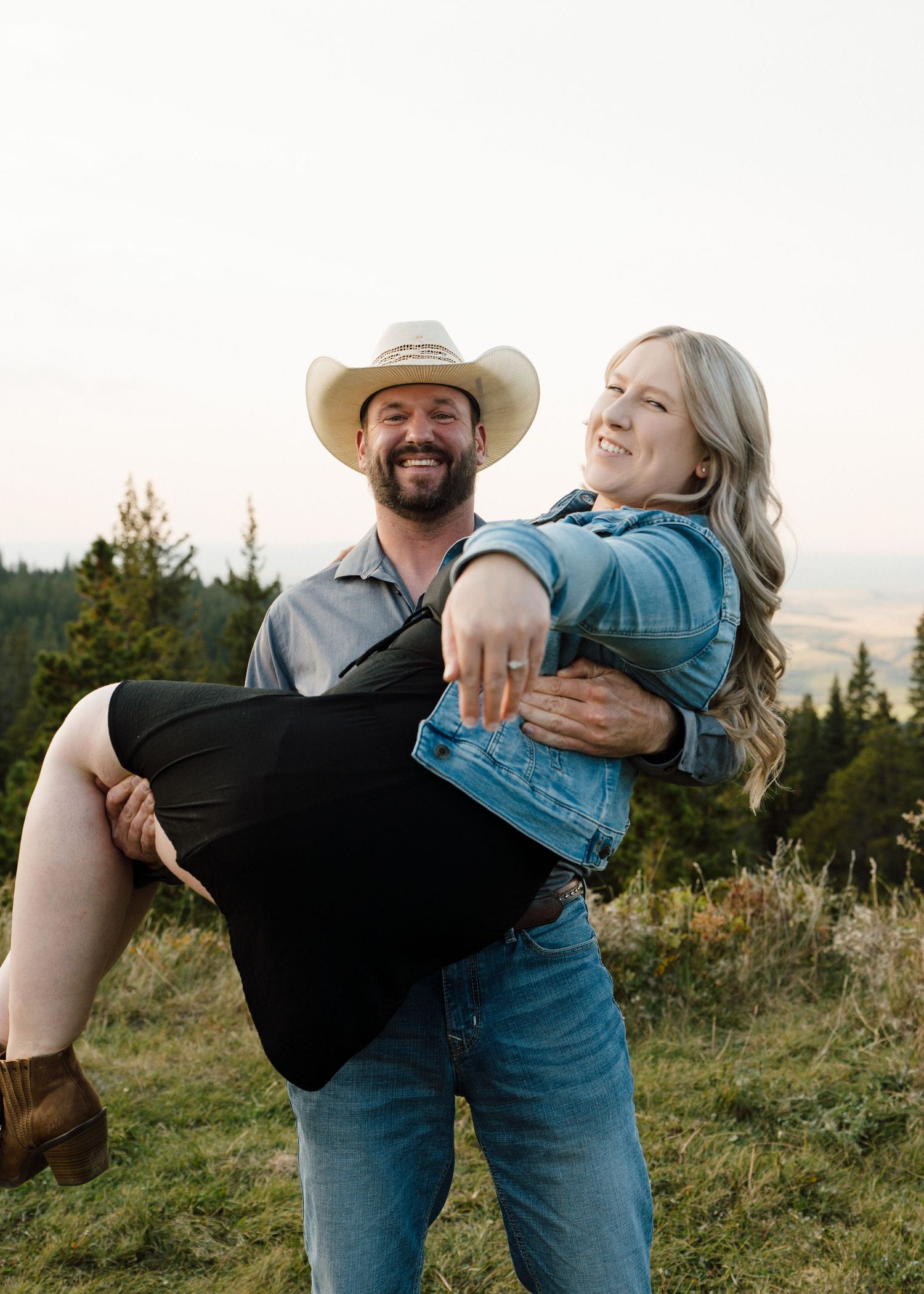 Smiling man in cowboy hat lifts laughing woman in black dress outdoors on hillside.