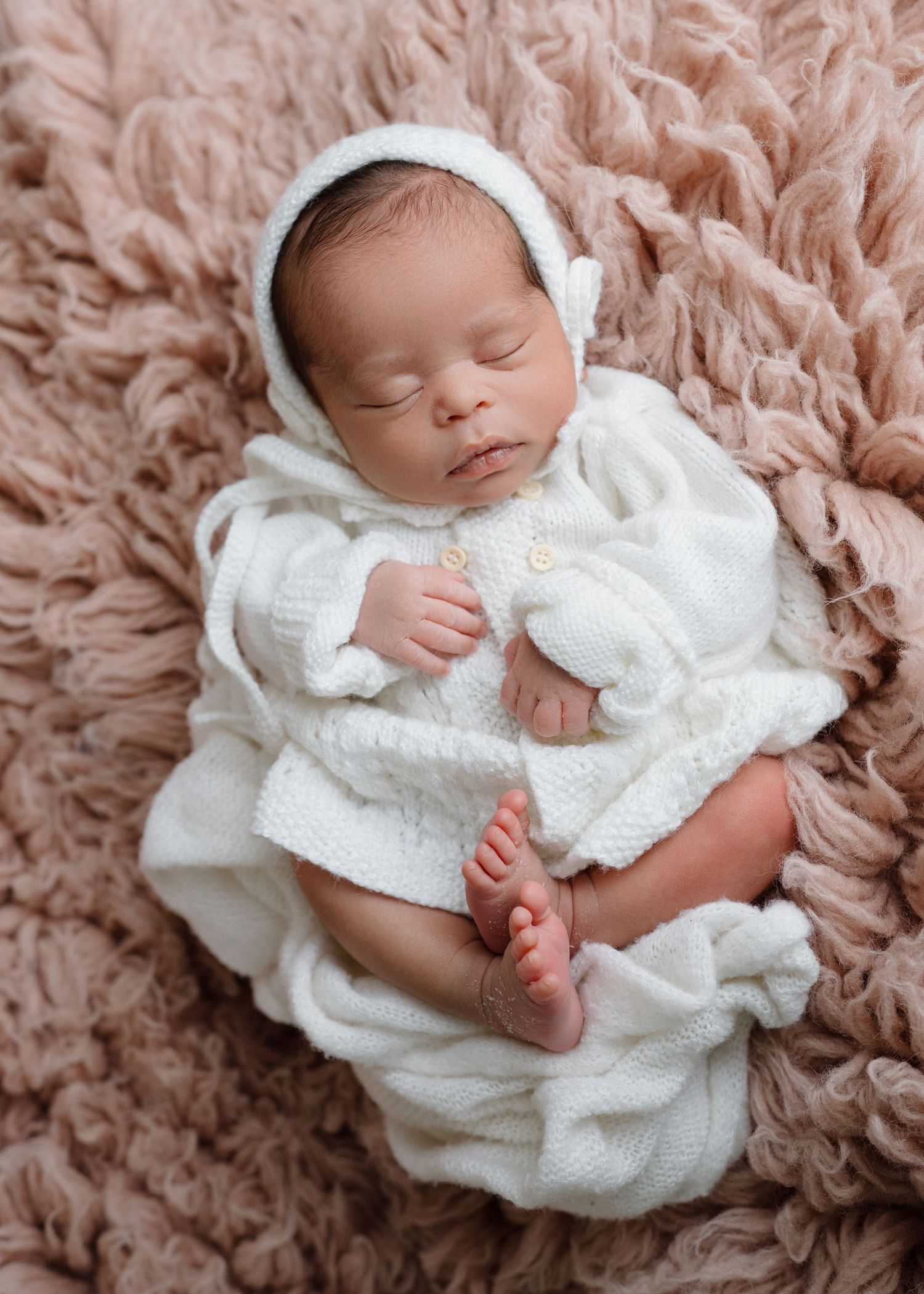 Peaceful newborn baby sleeping in white knit outfit and bonnet on soft pink fluffy blanket.