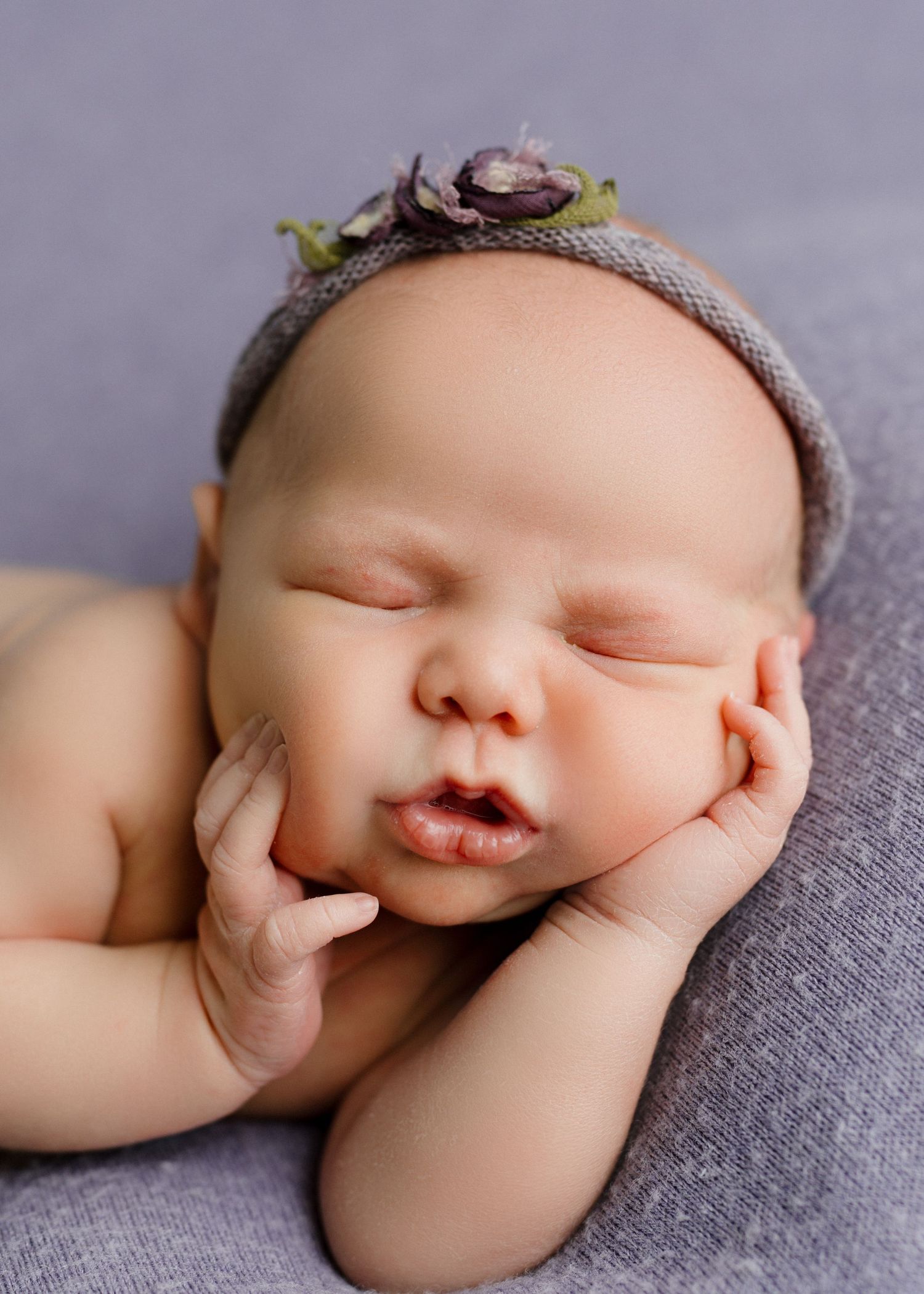 Sleeping newborn baby girl wearing a delicate purple floral headband on soft gray backdrop.