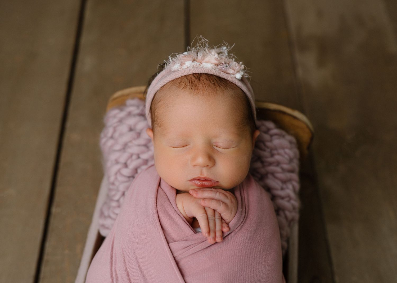Swaddled newborn baby girl in pink wrap sleeping in wooden bowl wearing a feathered pink crown headband.