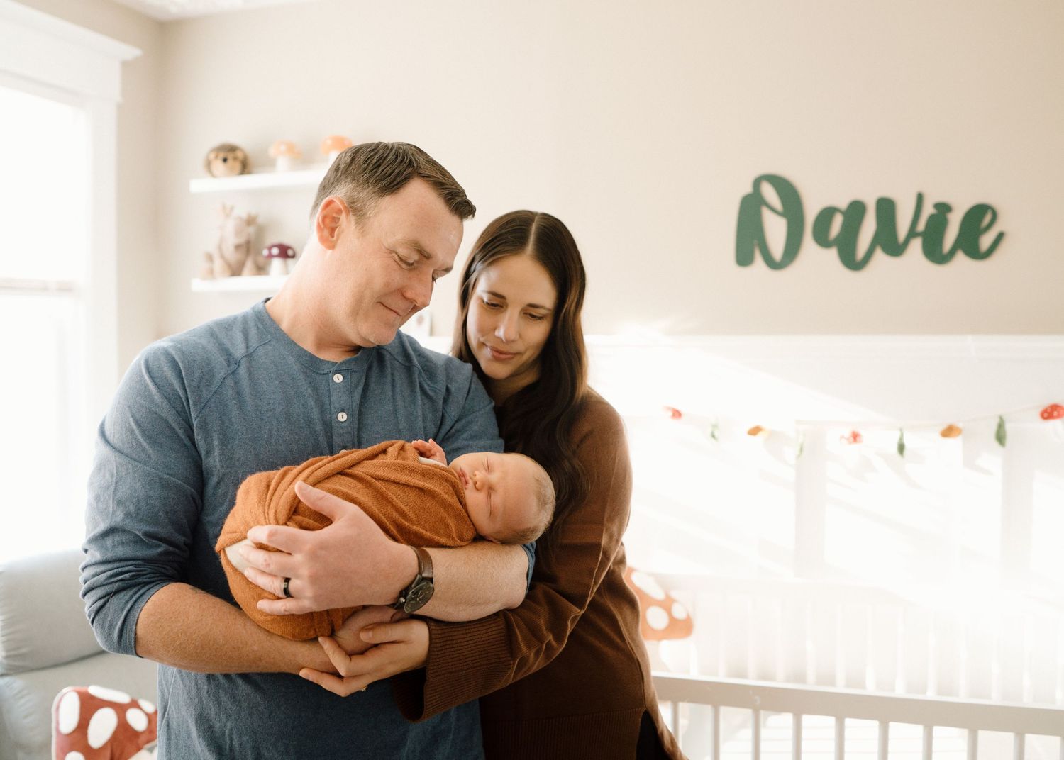 New parents cradle their swaddled newborn in a cozy nursery with a green name sign reading Davie on the wall.