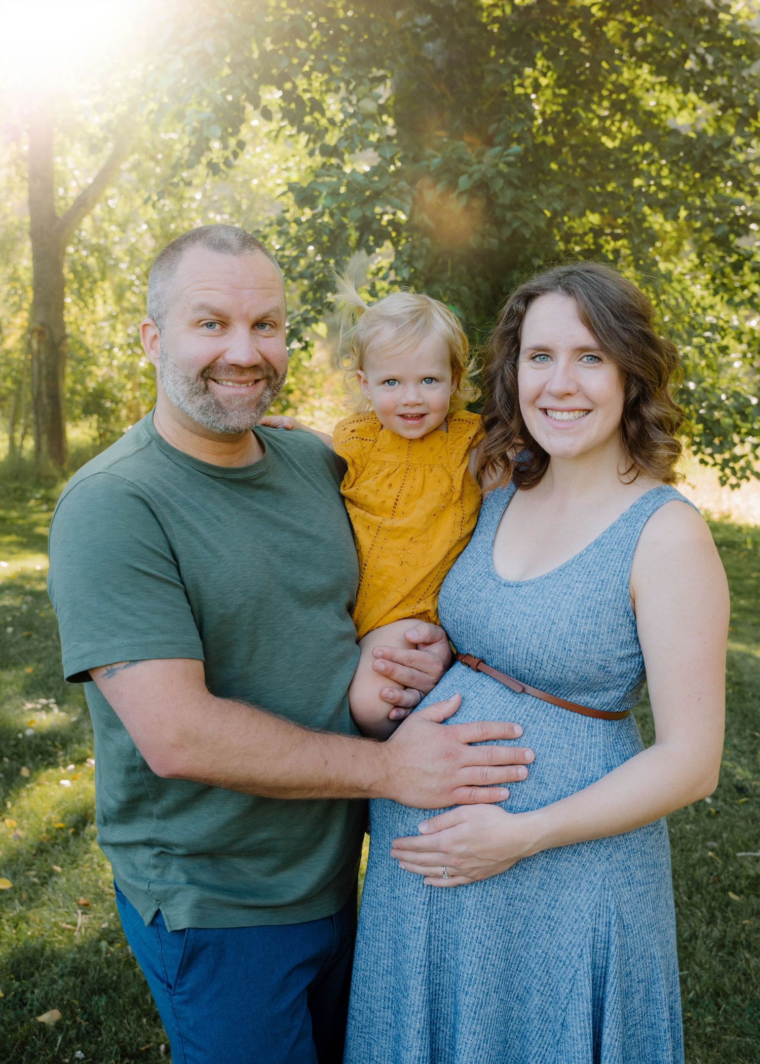 Expecting couple smiling in golden sunlight park setting, holding toddler girl in yellow romper during maternity session.