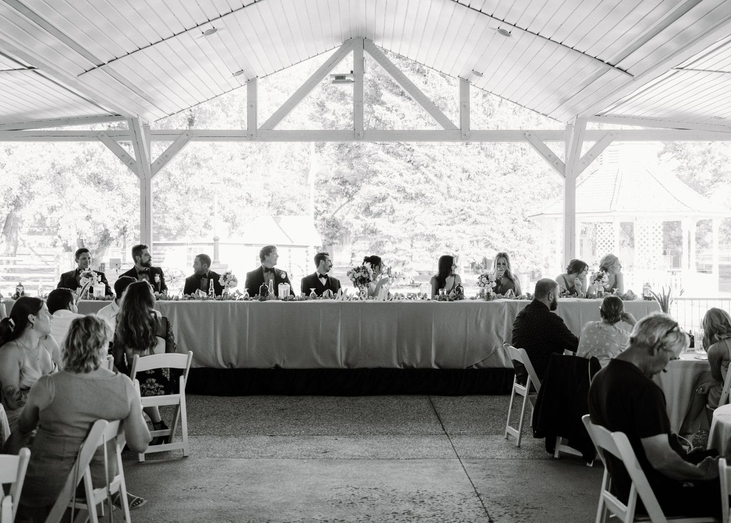 Wedding guests seated at a long reception table under an outdoor pavilion in black and white.
