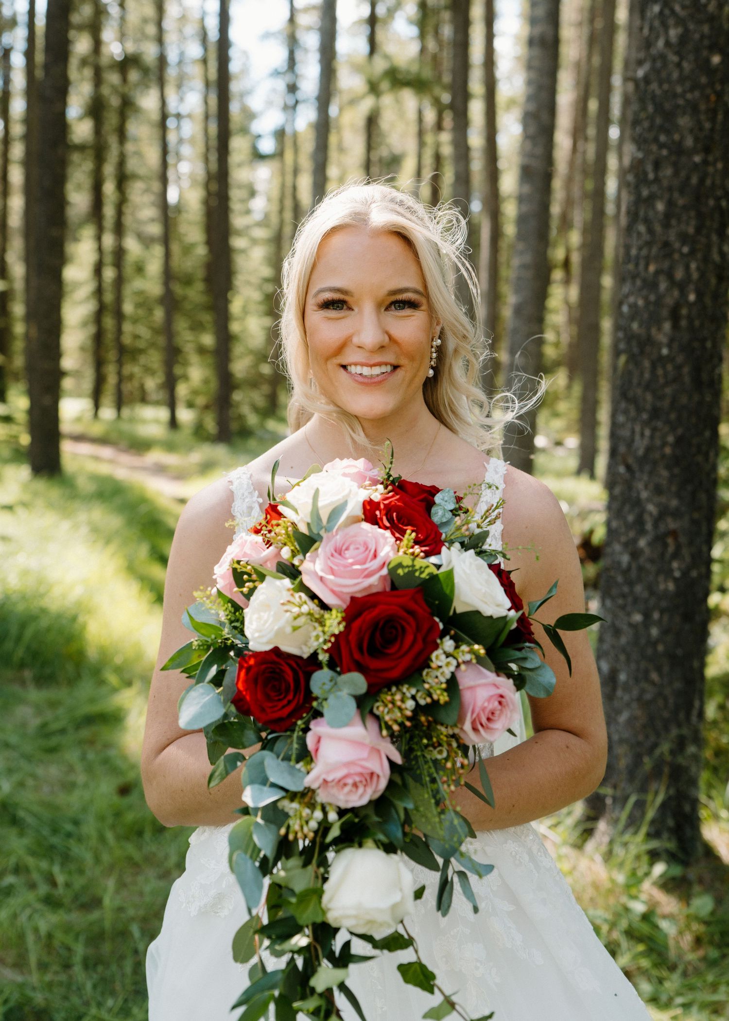 Smiling bride in white dress holding red and pink rose bouquet in a lush forest setting.