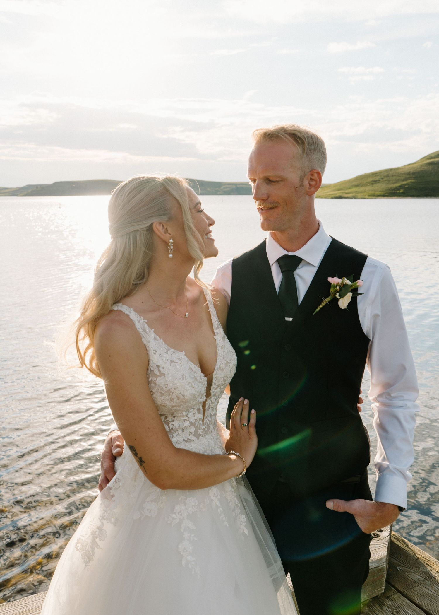 A bride and groom share a romantic moment by a lakeside at golden hour, dressed in wedding attire.