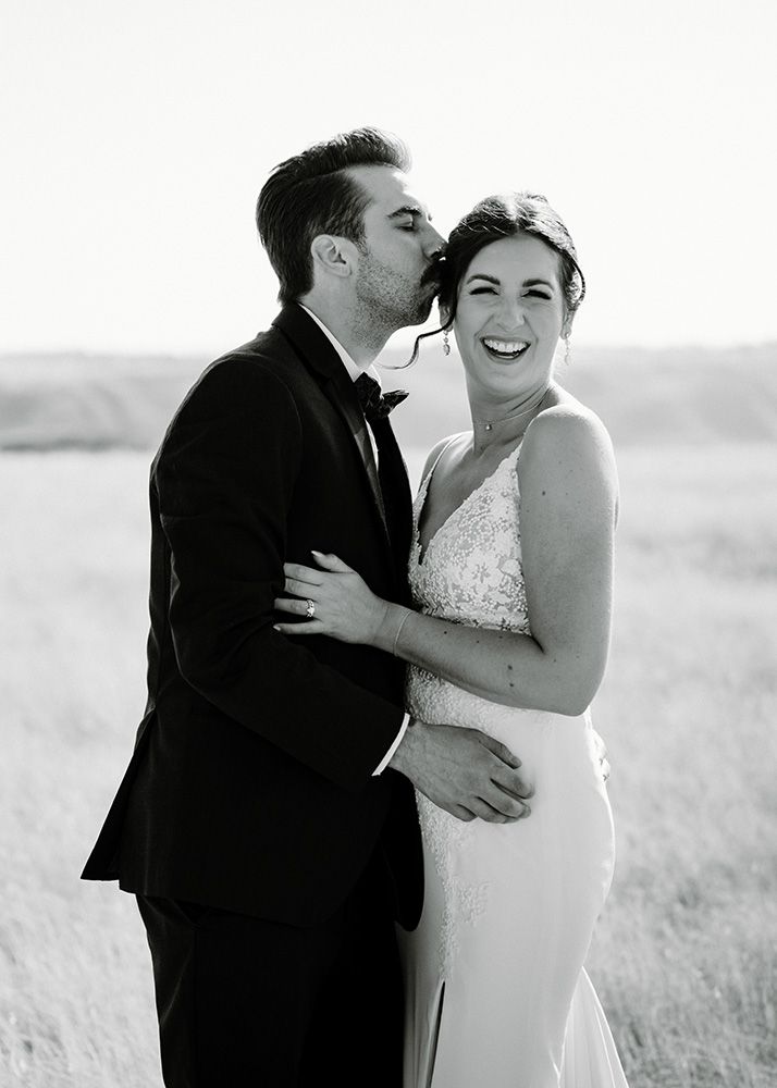 A happy bride laughs as groom kisses her cheek in a black and white wedding portrait in an open field.