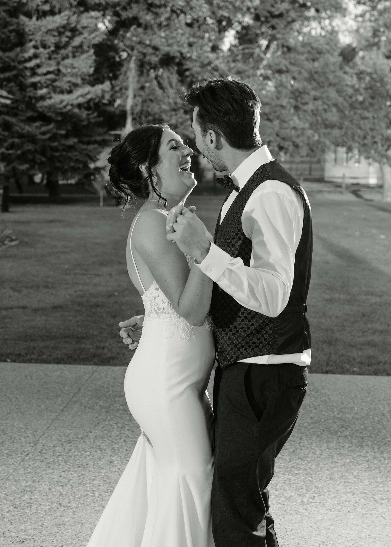 Black and white photo of a laughing bride in a mermaid gown dancing with a groom in a vest and bow tie outdoors.