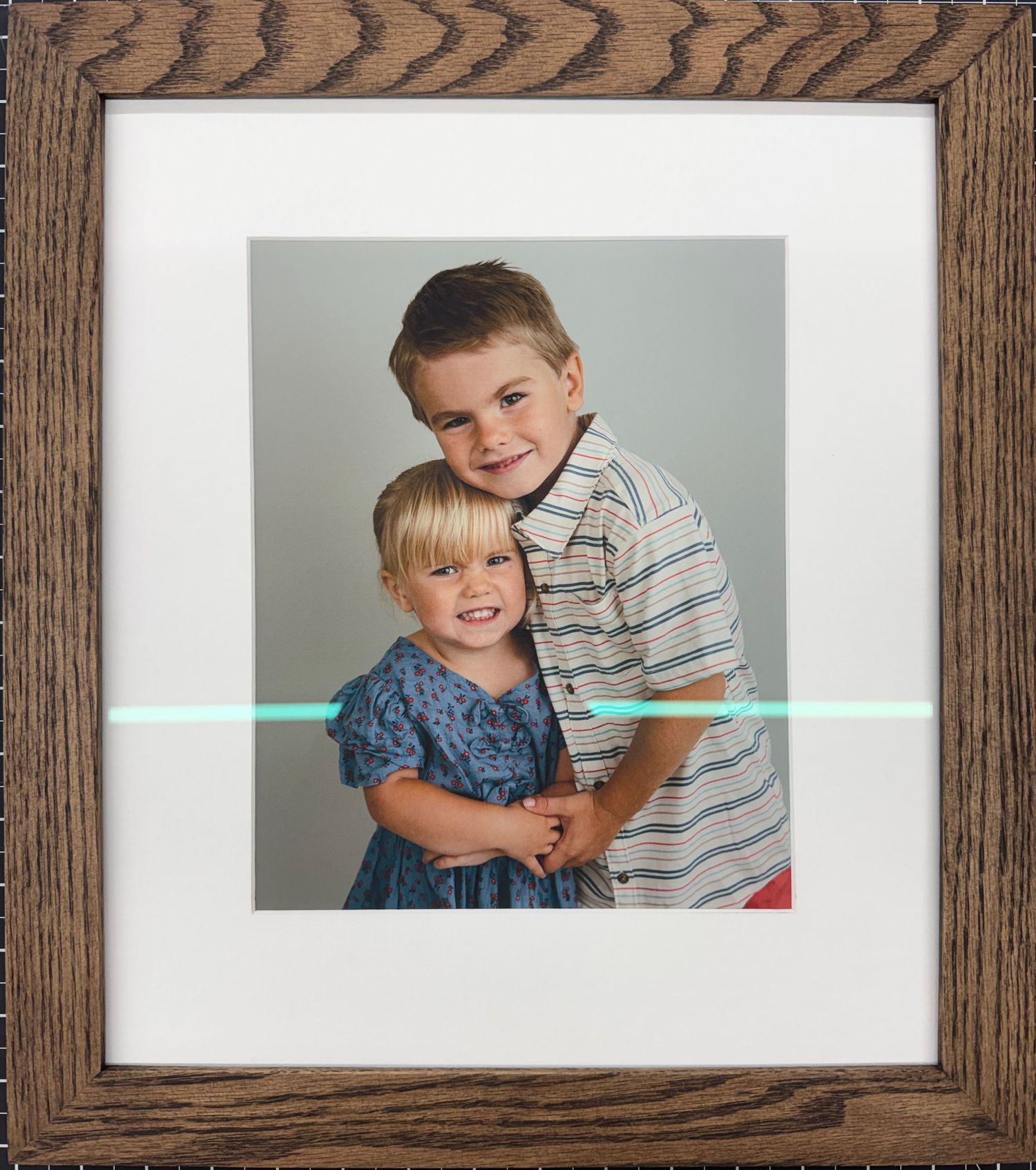 Framed portrait of two young siblings, a smiling boy in striped shirt hugging his little sister in a blue dress.