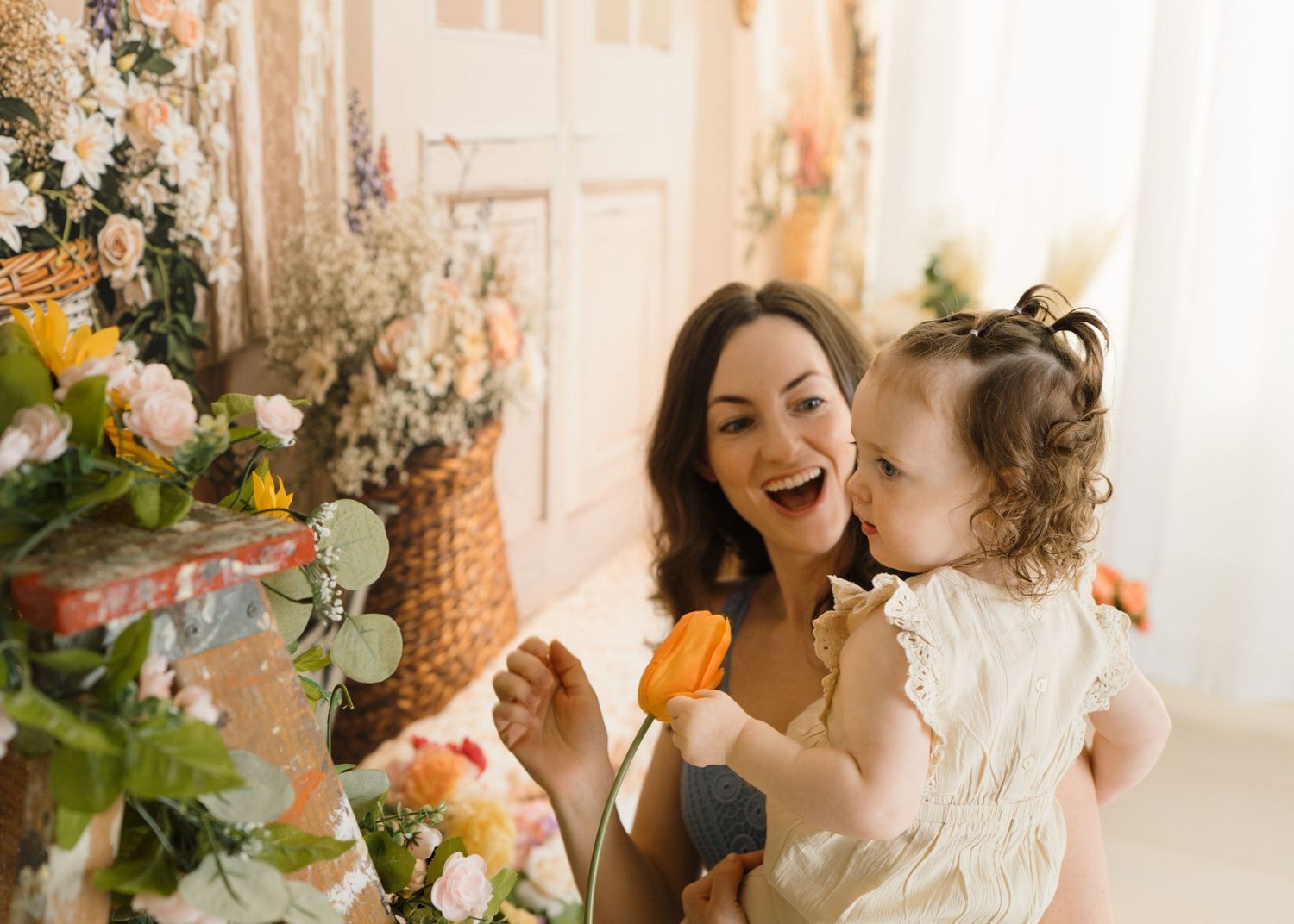 Laughing mother holds toddler daughter holding orange tulip in a bright floral decorated room.
