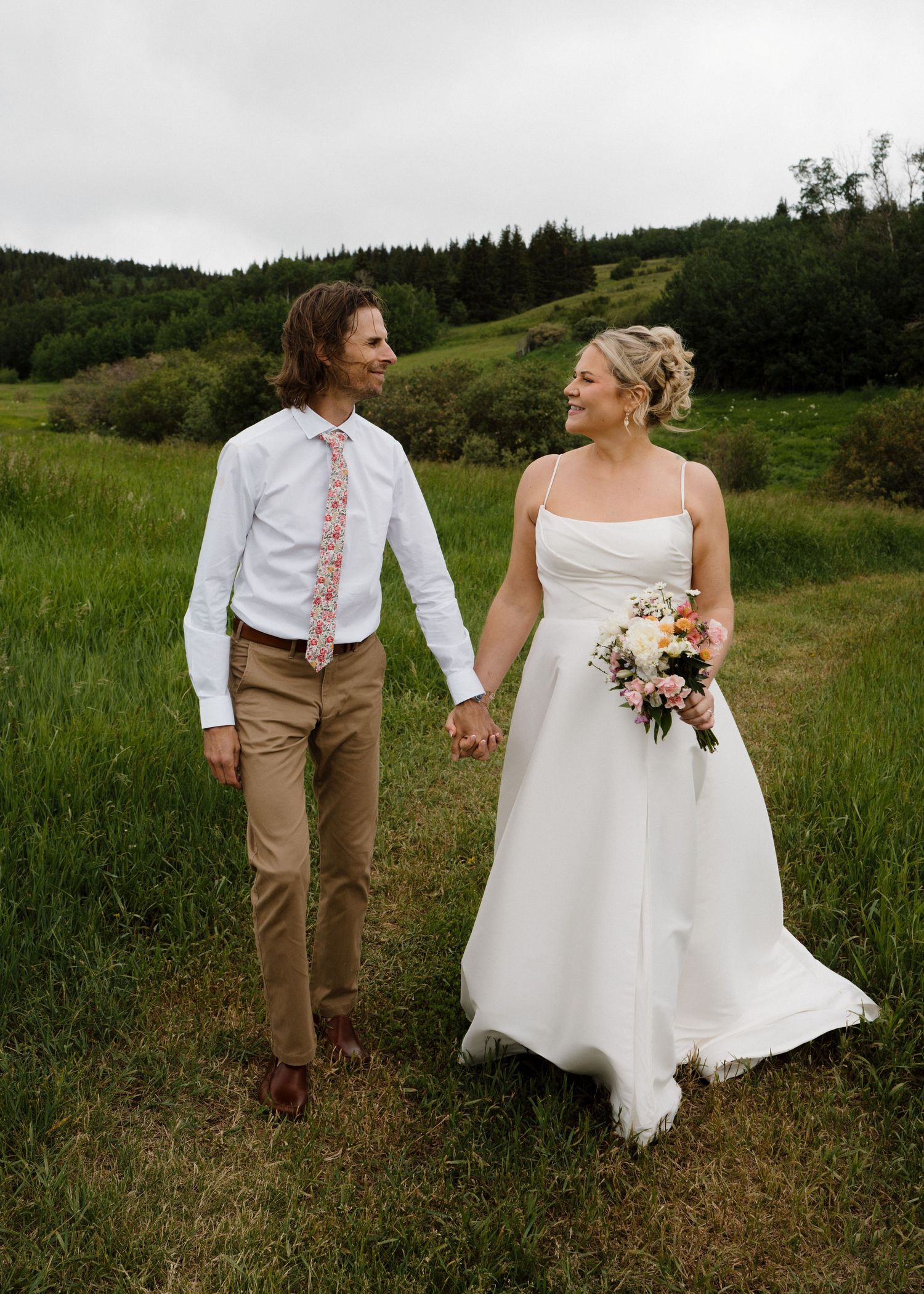 Bride in white spaghetti strap gown and groom in khakis walking hand in hand through a meadow.
