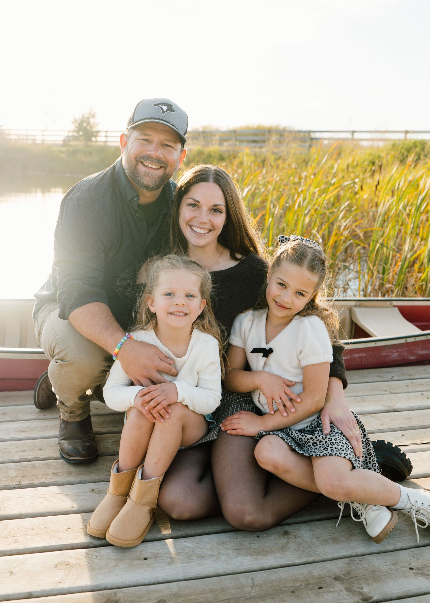 Happy family of four posing on a wooden dock near a lake and tall grass during golden hour.