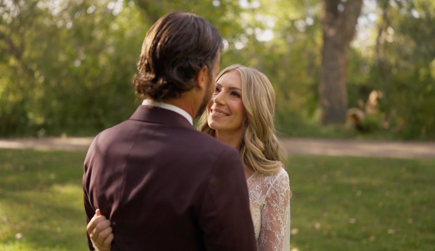 Couple sharing a romantic moment outdoors at a wedding, surrounded by lush green trees in soft natural light.