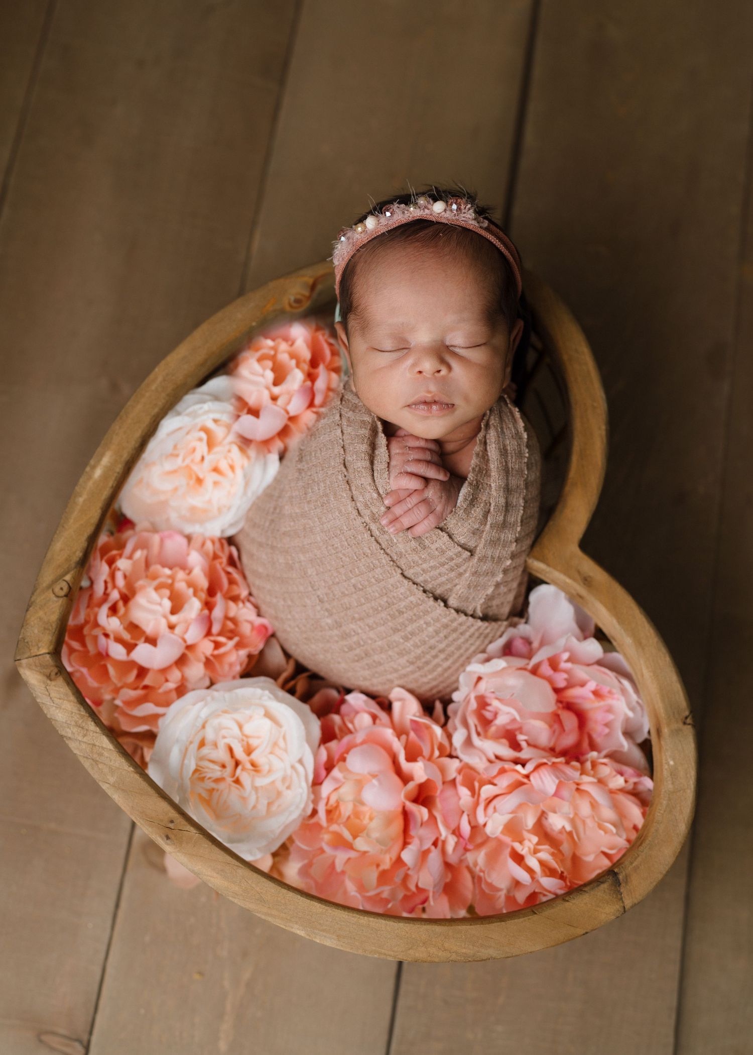 Sleeping swaddled newborn baby in heart-shaped wooden bowl surrounded by pink and white flowers.