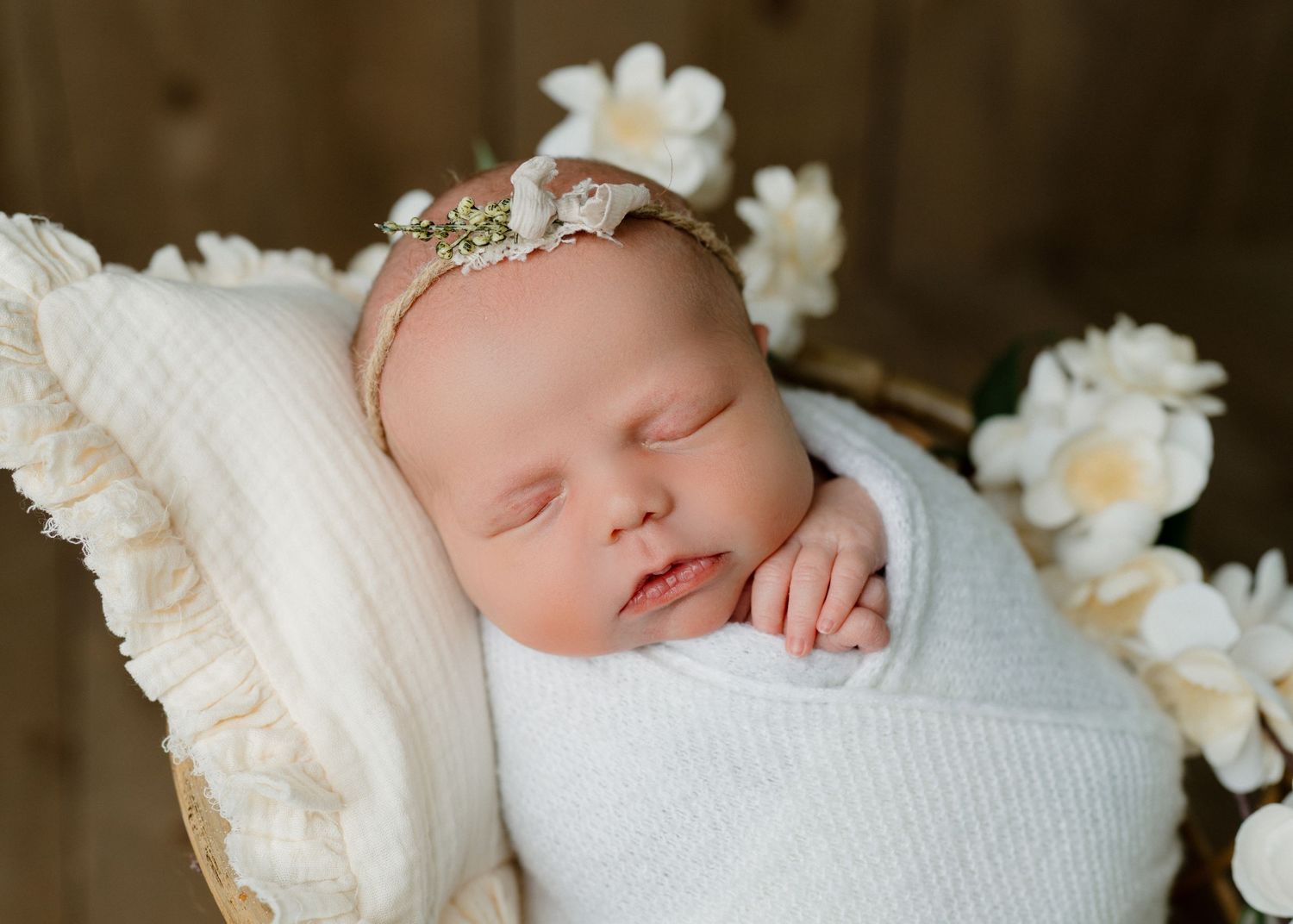 Peaceful newborn baby sleeping in white swaddle with floral headband surrounded by white flowers.