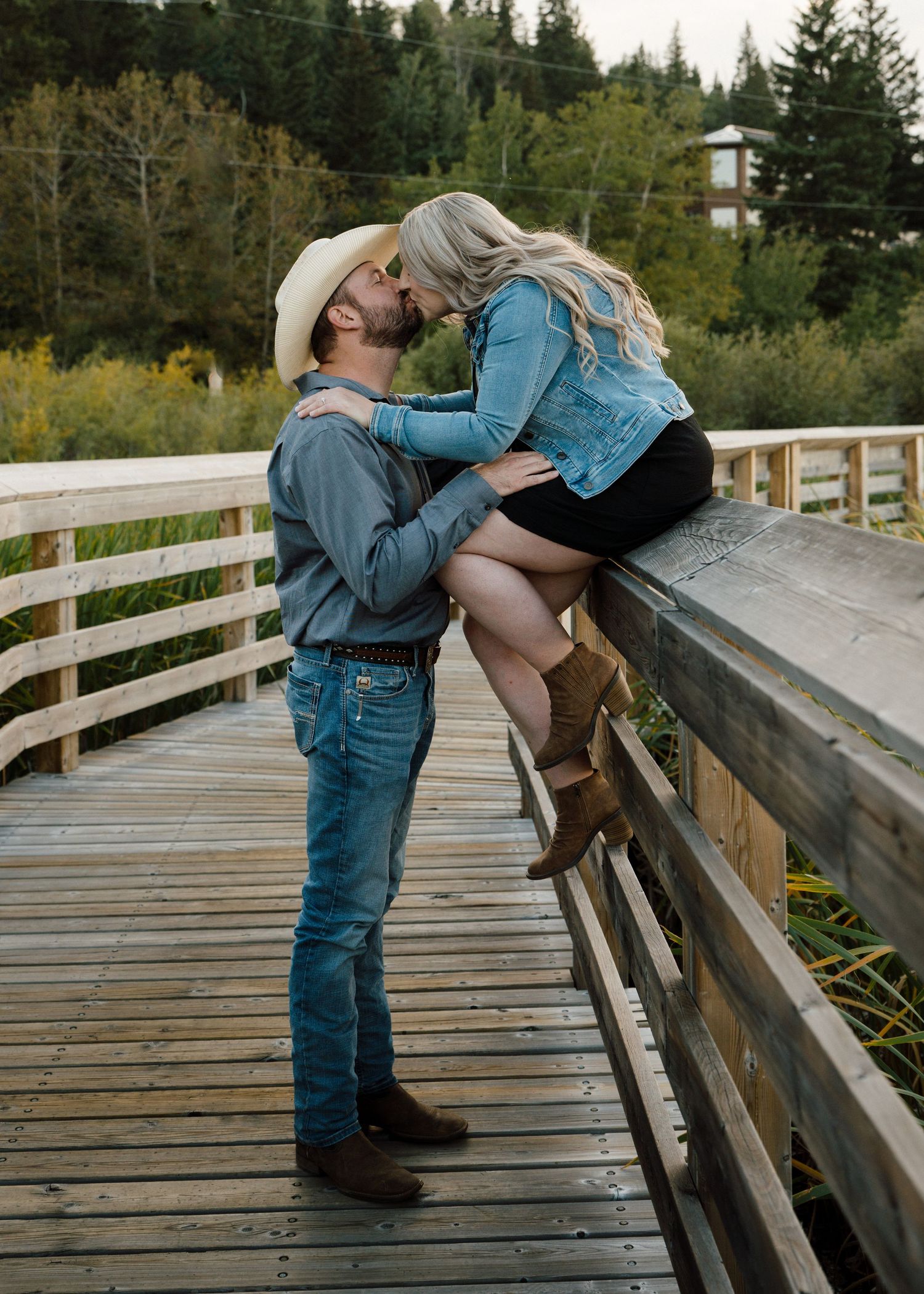 Couple kissing romantically on wooden bridge surrounded by lush green trees.
