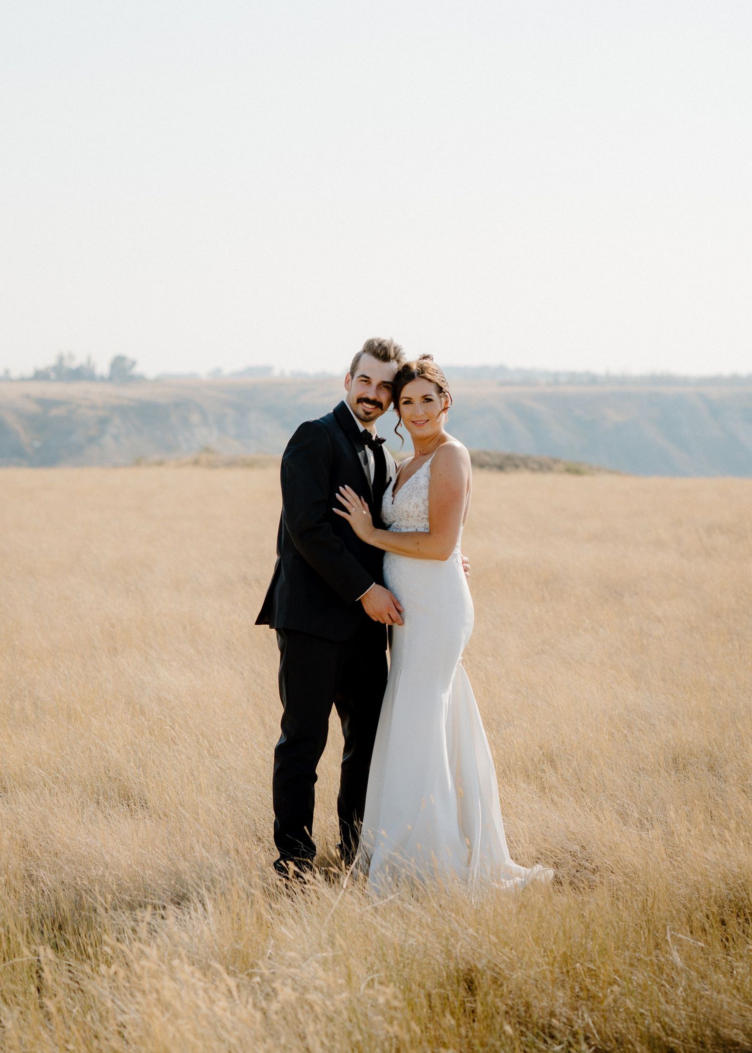 Bride and groom pose together in a golden wheat field with rolling hills in the background.