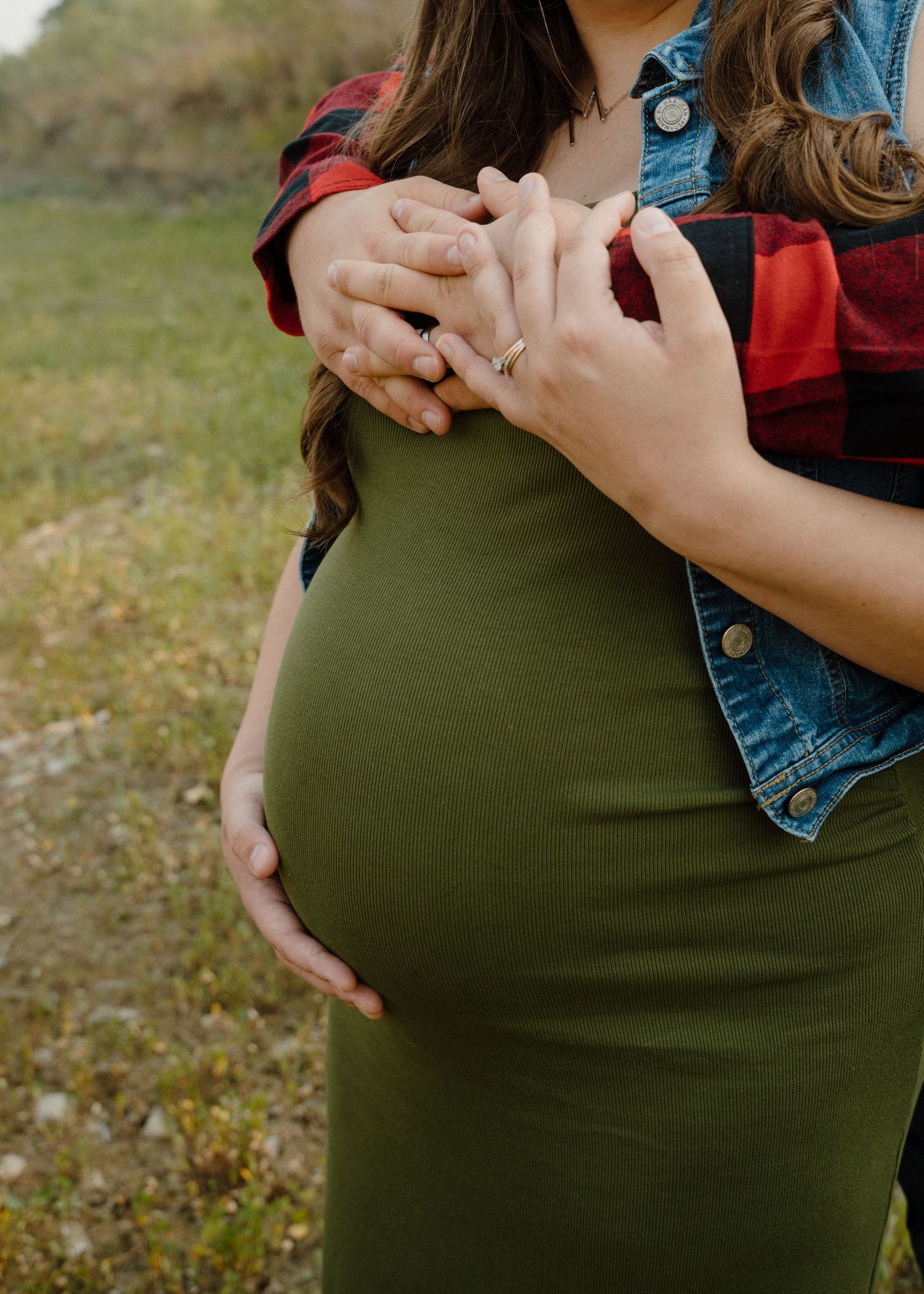 Close-up of pregnant woman in olive green dress with partner's hands forming heart shape over baby bump outdoors.