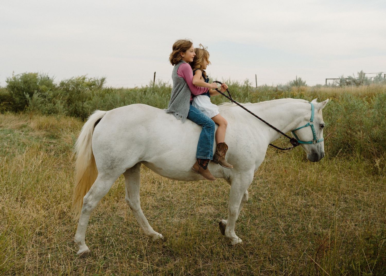 Two children riding together on a white horse through a grassy field on a cloudy day.