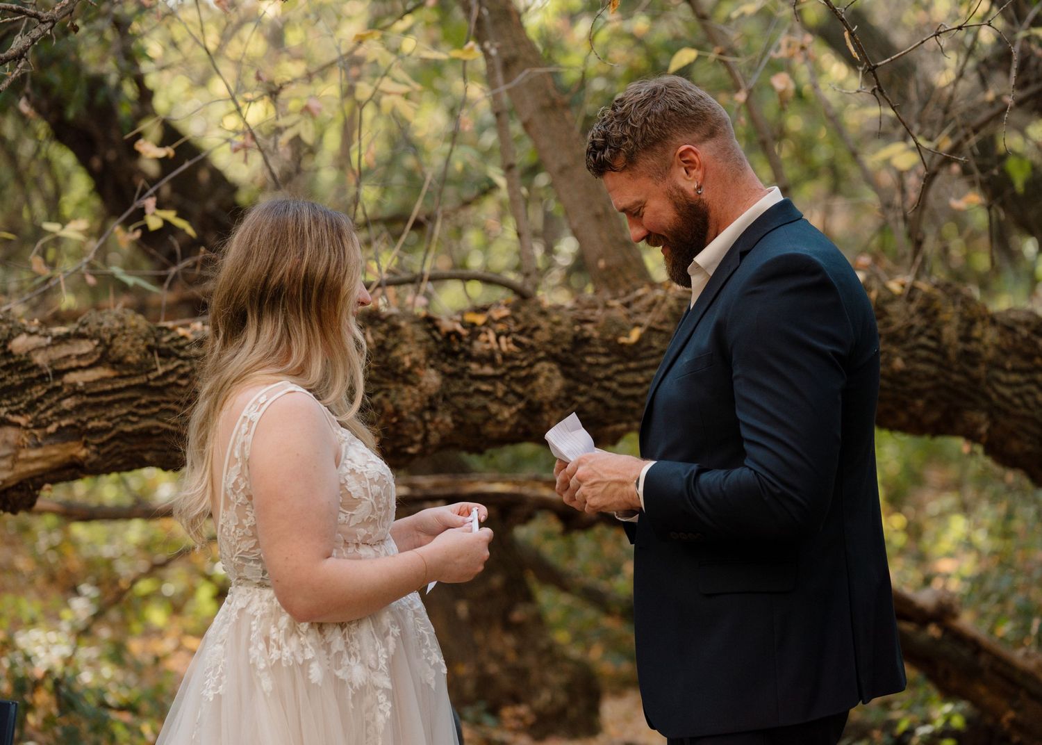 Groom in navy suit reading vows to bride in lace dress during an intimate outdoor woodland ceremony.