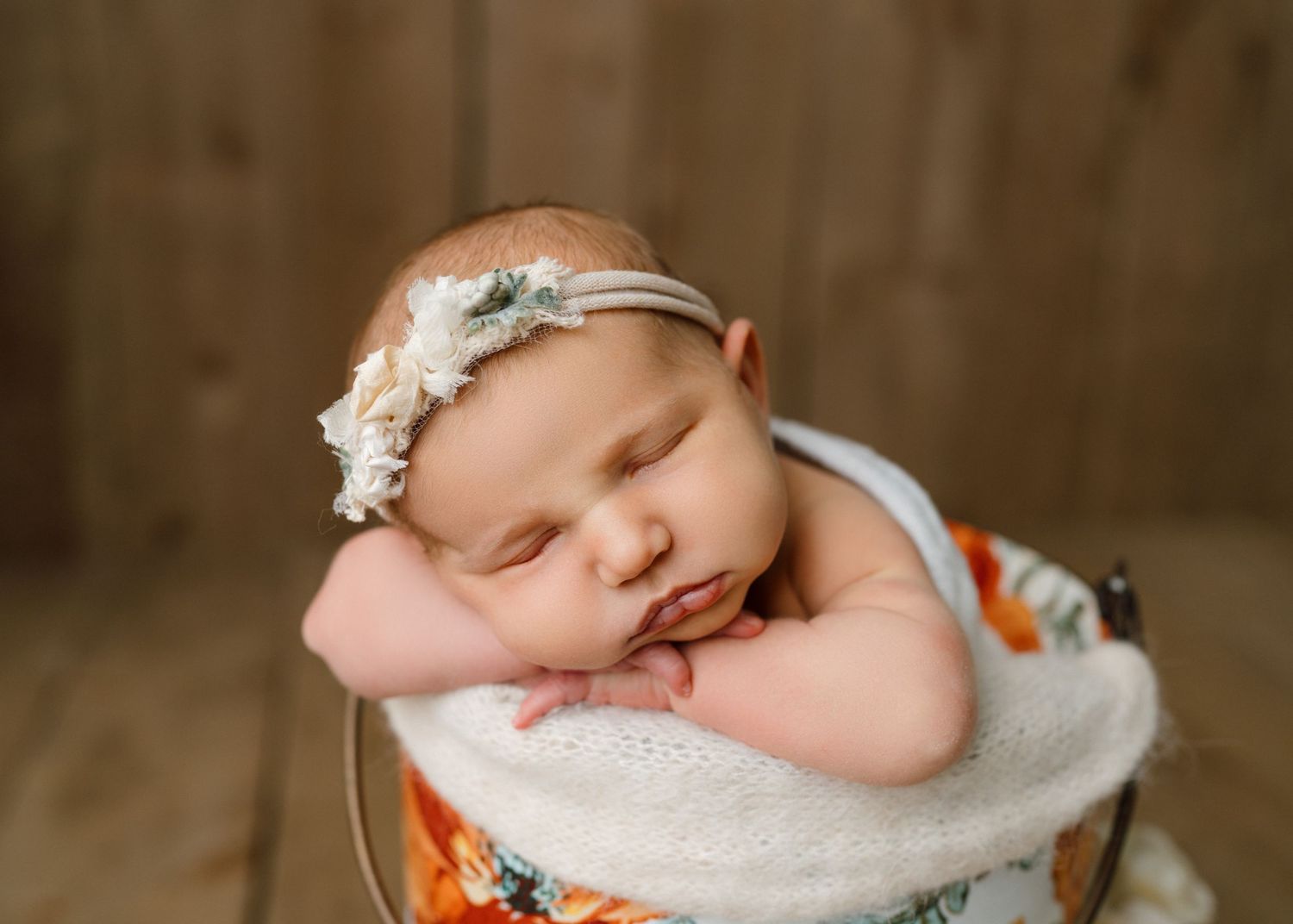 Sleeping newborn baby girl in white swaddle with floral headband resting in a decorative bucket.