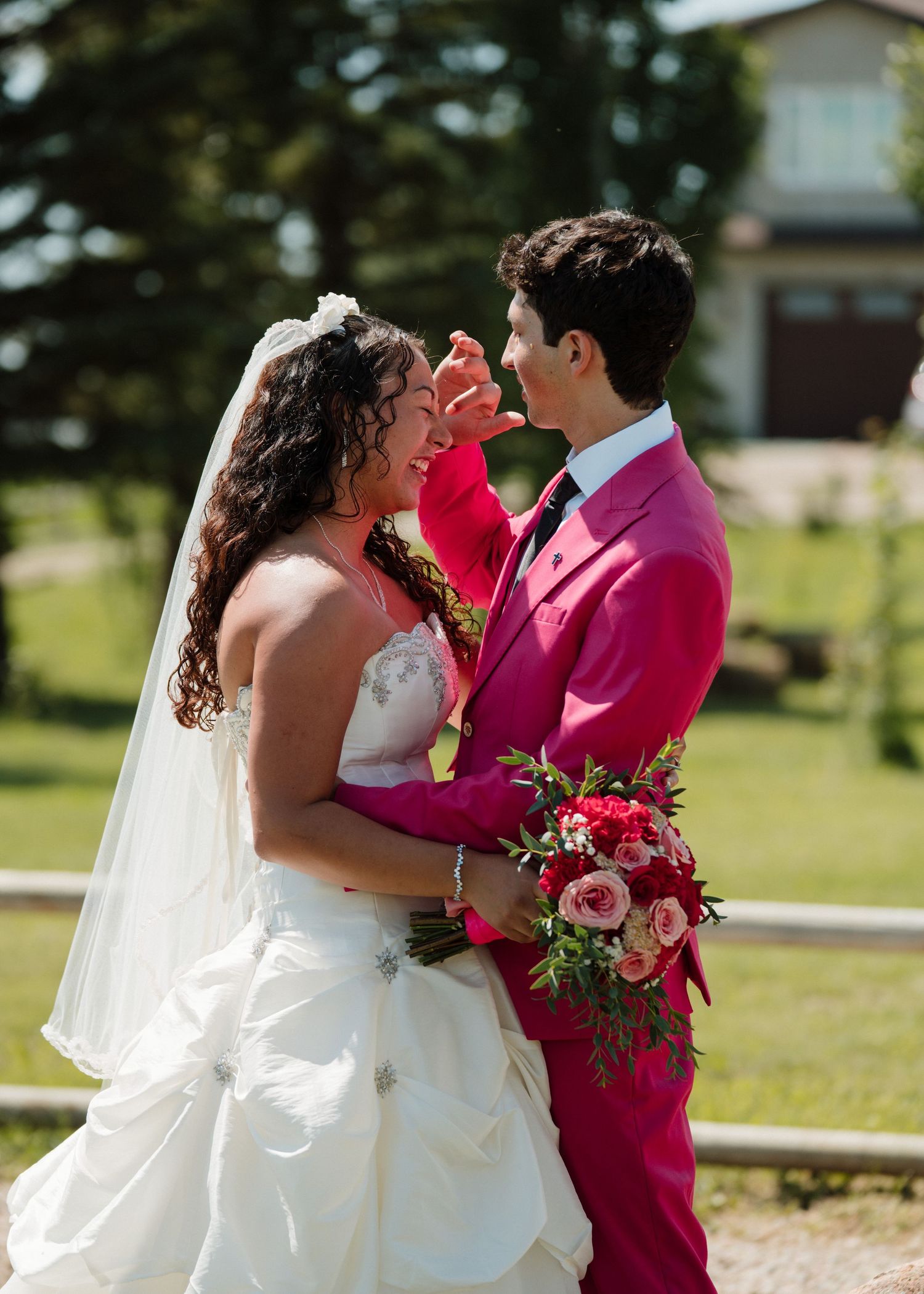 Bride and groom share tender moment, groom in pink suit wiping bride's tears outdoors.