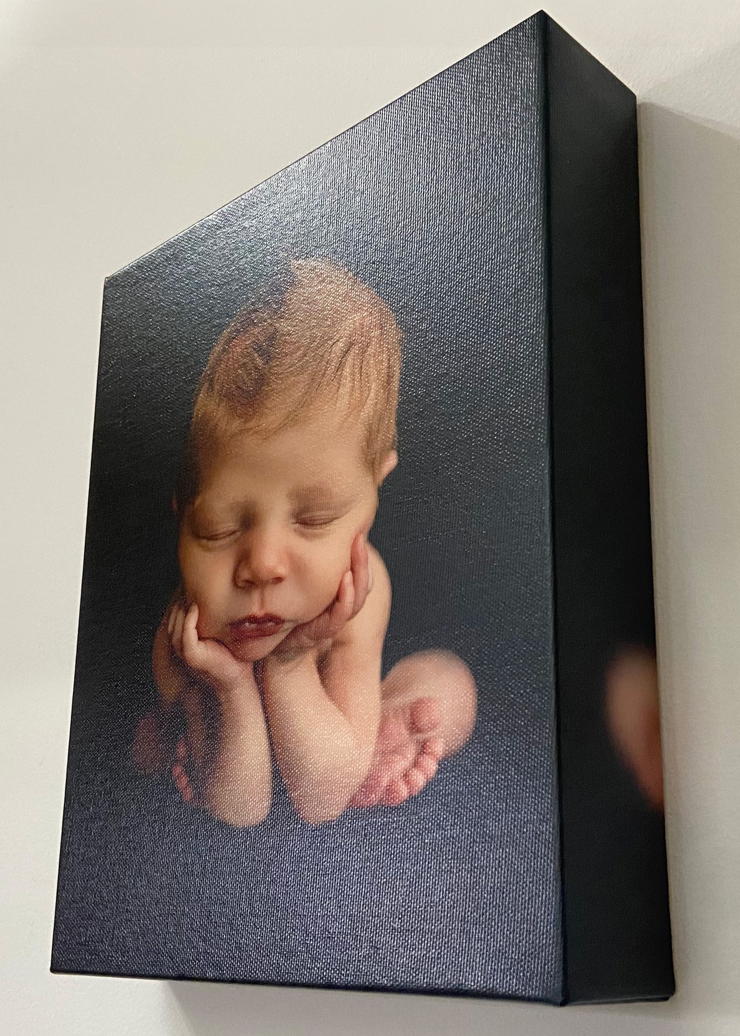 Side-angle view of a dark canvas print featuring a peaceful sleeping newborn baby resting chin on hands.
